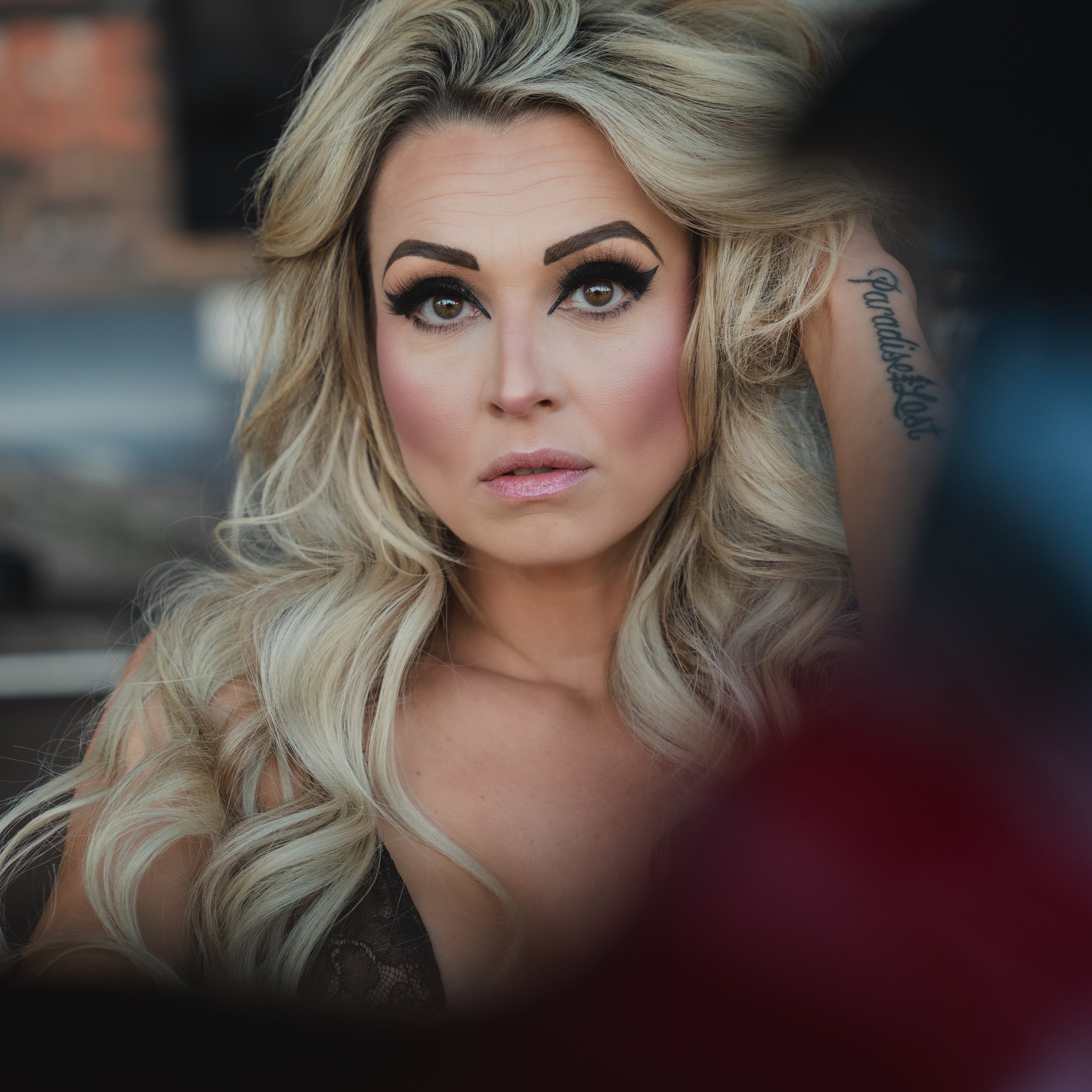 Close-up of a woman with blonde hair and makeup, holding her hair back with one hand, tattoo on her arm, and wearing a black lace top.