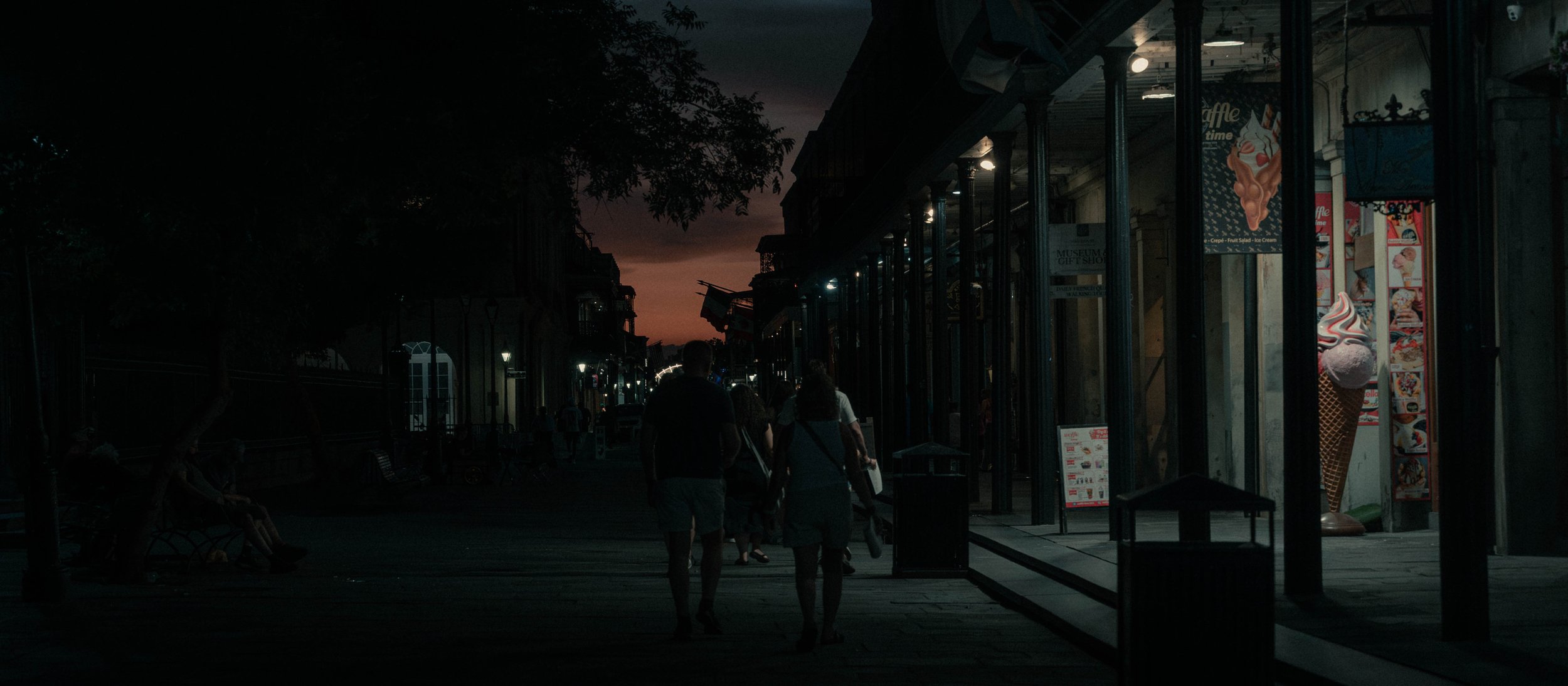 Nighttime street scene with a group of people walking past a store with ice cream advertisements, and a sunset sky in the background.
