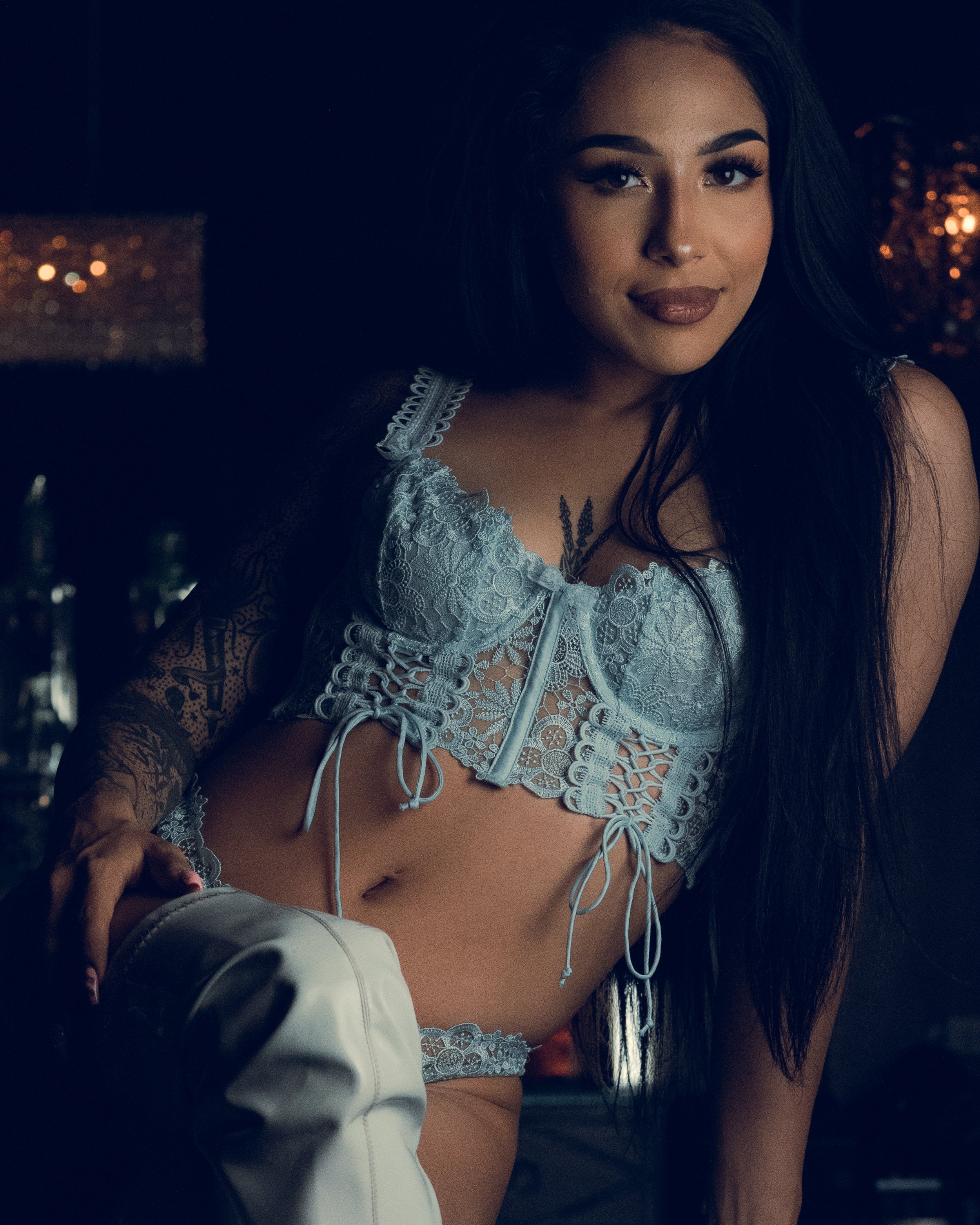 Close-up of a woman with dark hair, wearing a white lace top, smiling softly, with blurred city lights in the background at night.