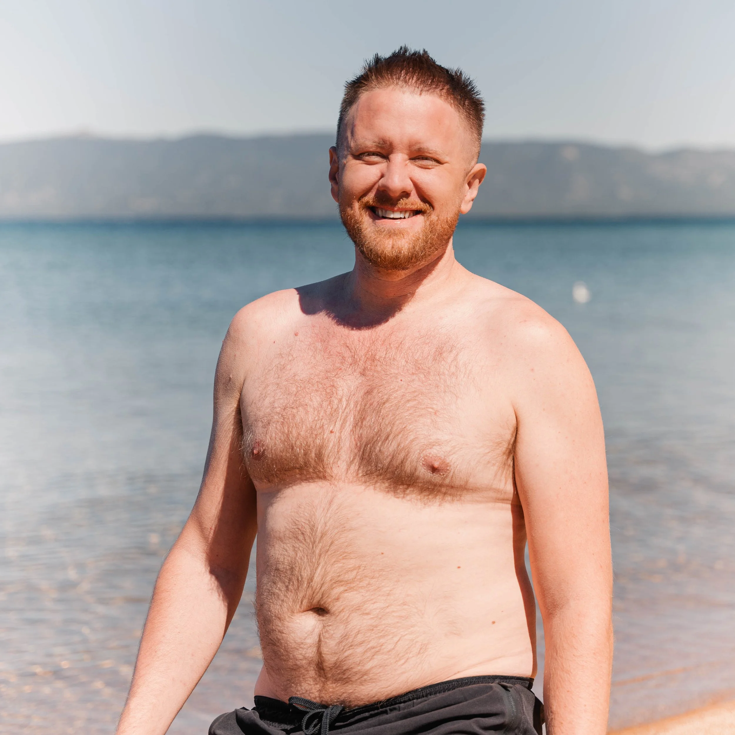 Close-up of a smiling man with short hair and a beard, shirtless, standing at the beach with water and distant mountains in the background.