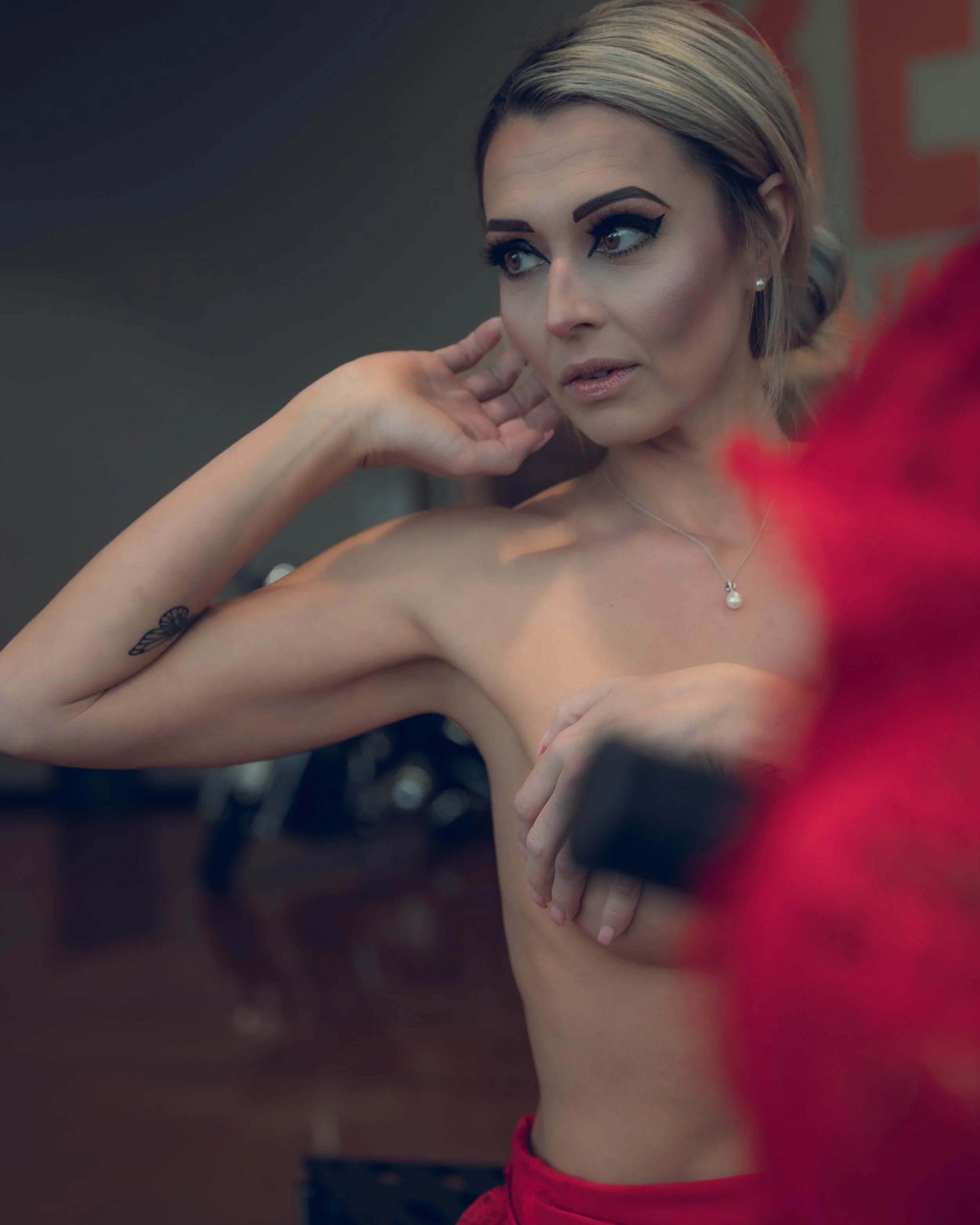 A woman with short blonde hair, bold eyeliner, and pearl earrings adjusting her hair near her ear, with her back partially visible and a blurred background.