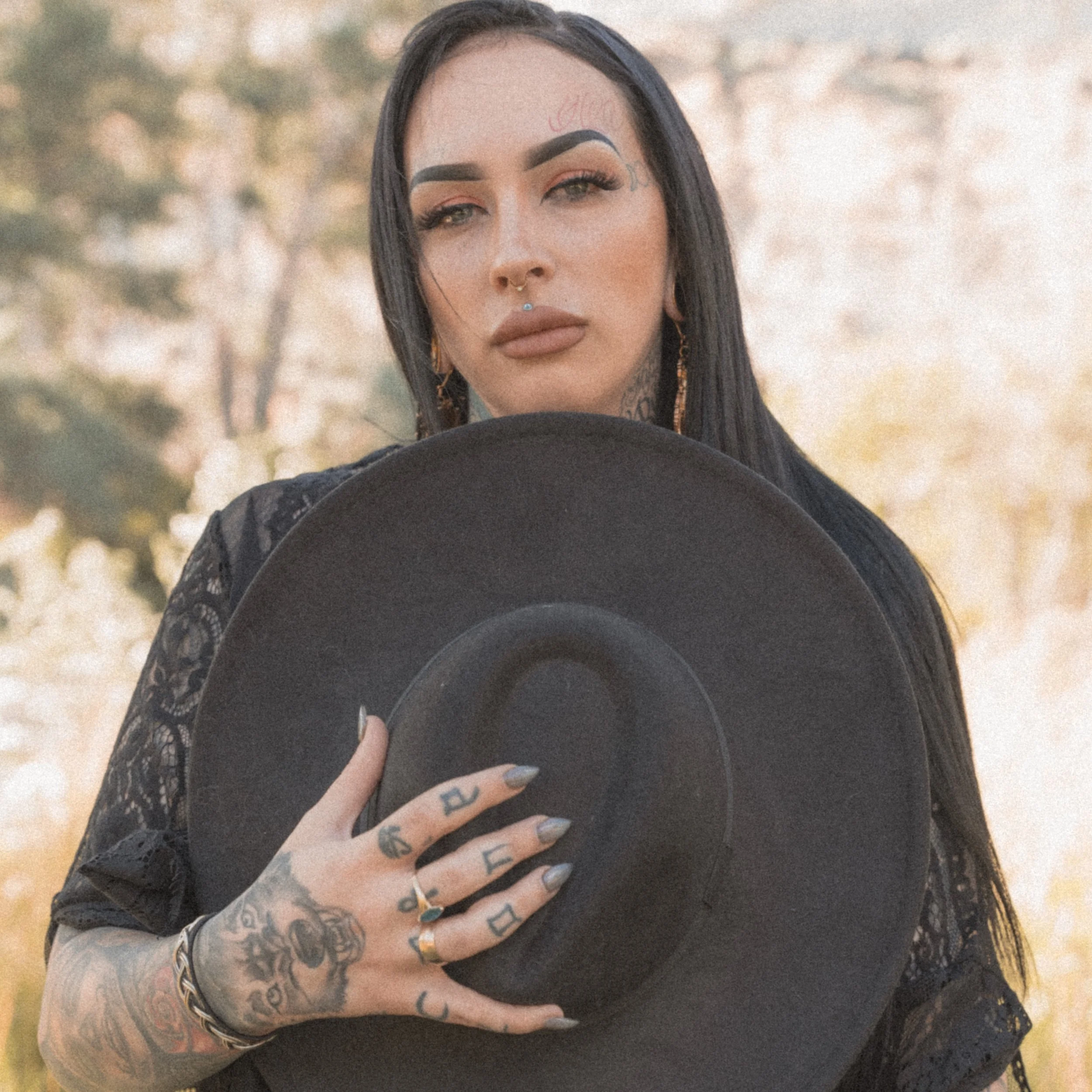 A young woman with dark hair and tattoos on her hand and fingers, holding a black hat in front of her face. She has bold makeup with dark eyebrows, pink eyeshadow, and piercings including a septum and a tongue piercing, outdoors with blurry trees in 