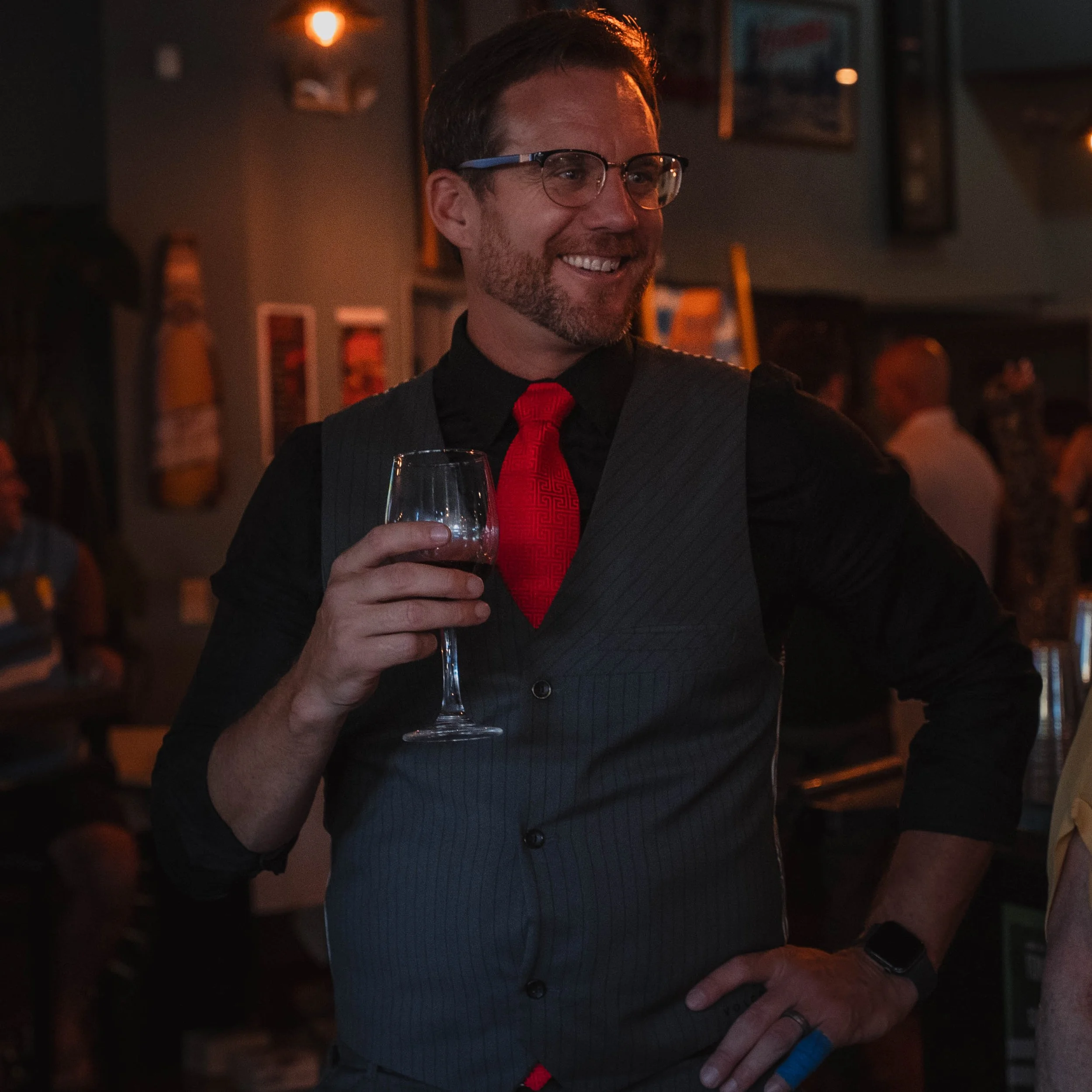 A smiling man wearing glasses, a gray vest, and a red tie at a social gathering or party, holding a glass of wine in a dimly-lit environment with warm lighting.