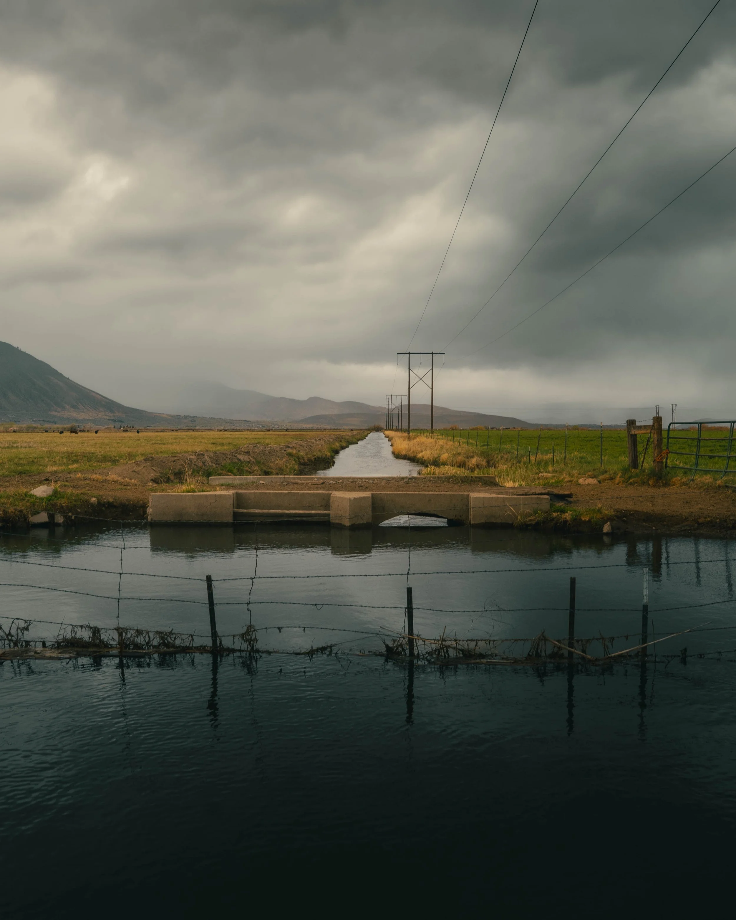 Dark stormy clouds over a rural landscape with mountains in the distance, power lines running through a field, and a water canal in the foreground.