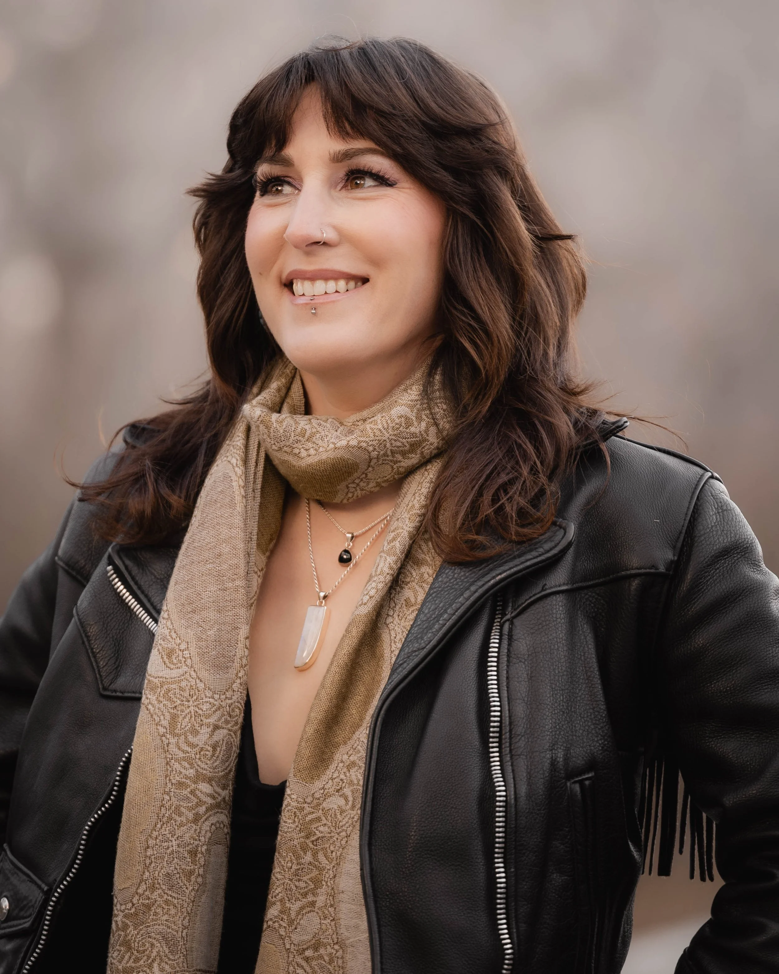 A woman with dark brown, wavy hair and light skin, smiling while wearing a black leather jacket, beige patterned scarf, and layered jewelry, outdoors with a blurred background.