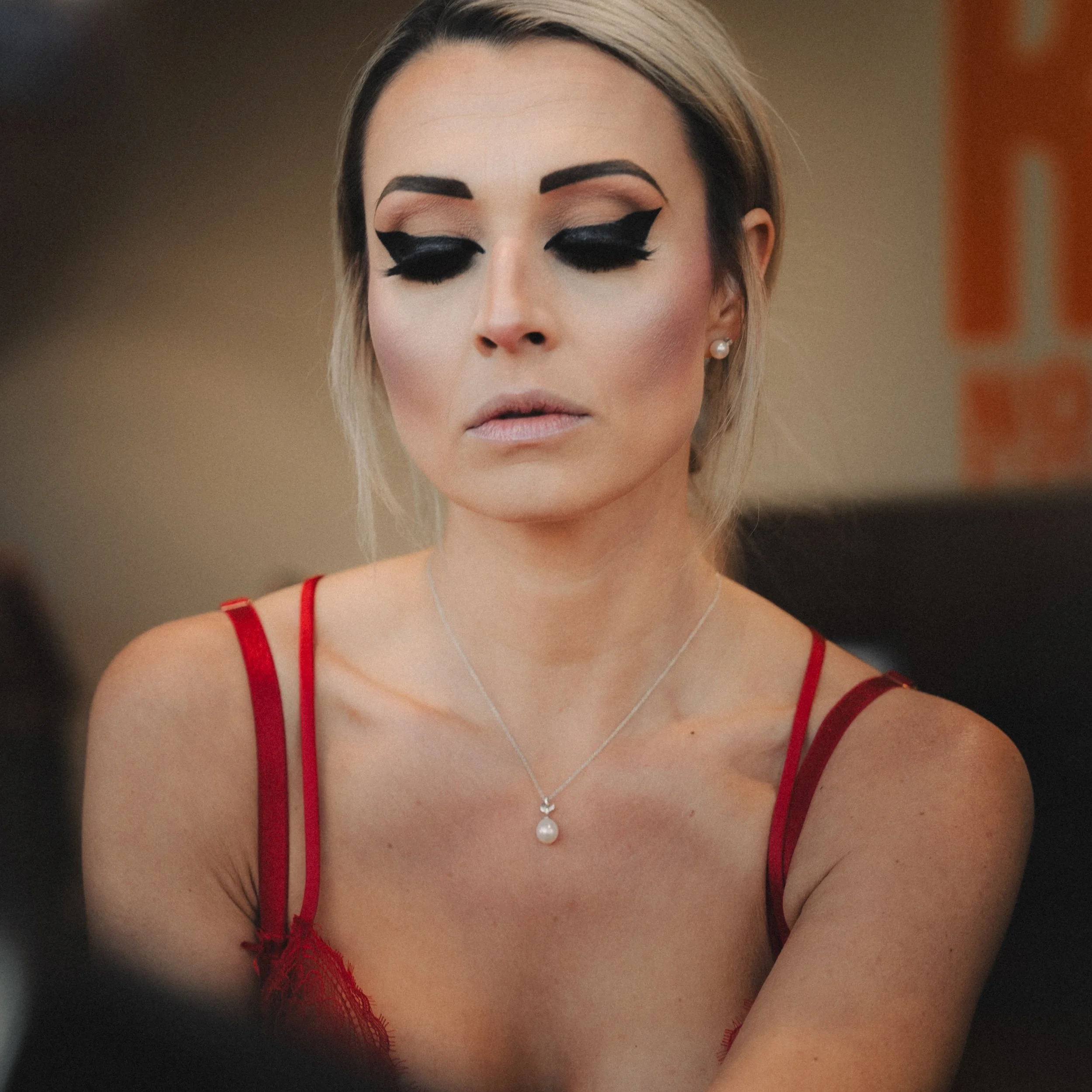 A woman with closed eyes wearing dramatic black eyeliner, pearl earrings, a red lace top, and a pearl pendant necklace.