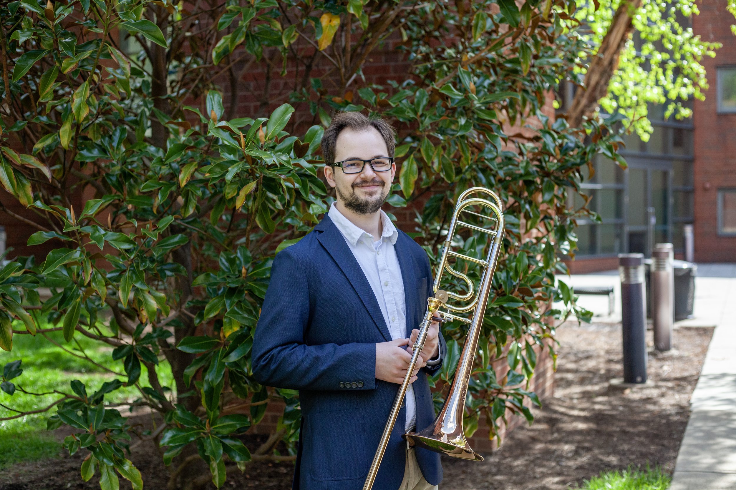 A man in a navy suit and glasses holding a trombone outdoors in front of green leafy bushes and a red brick building.