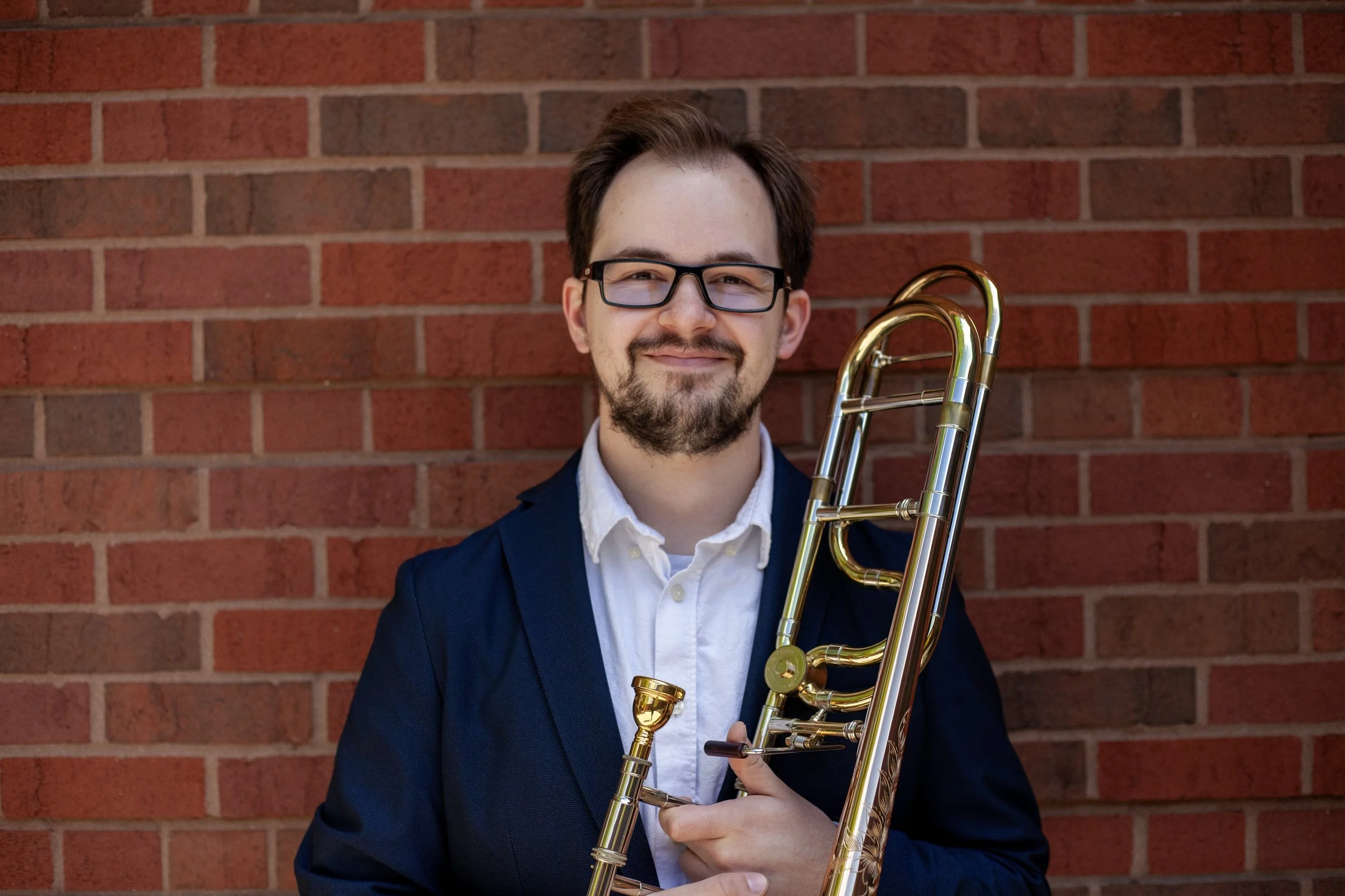 A young man with glasses, a beard, and dark hair holding a brass trombone, standing in front of a red brick wall.
