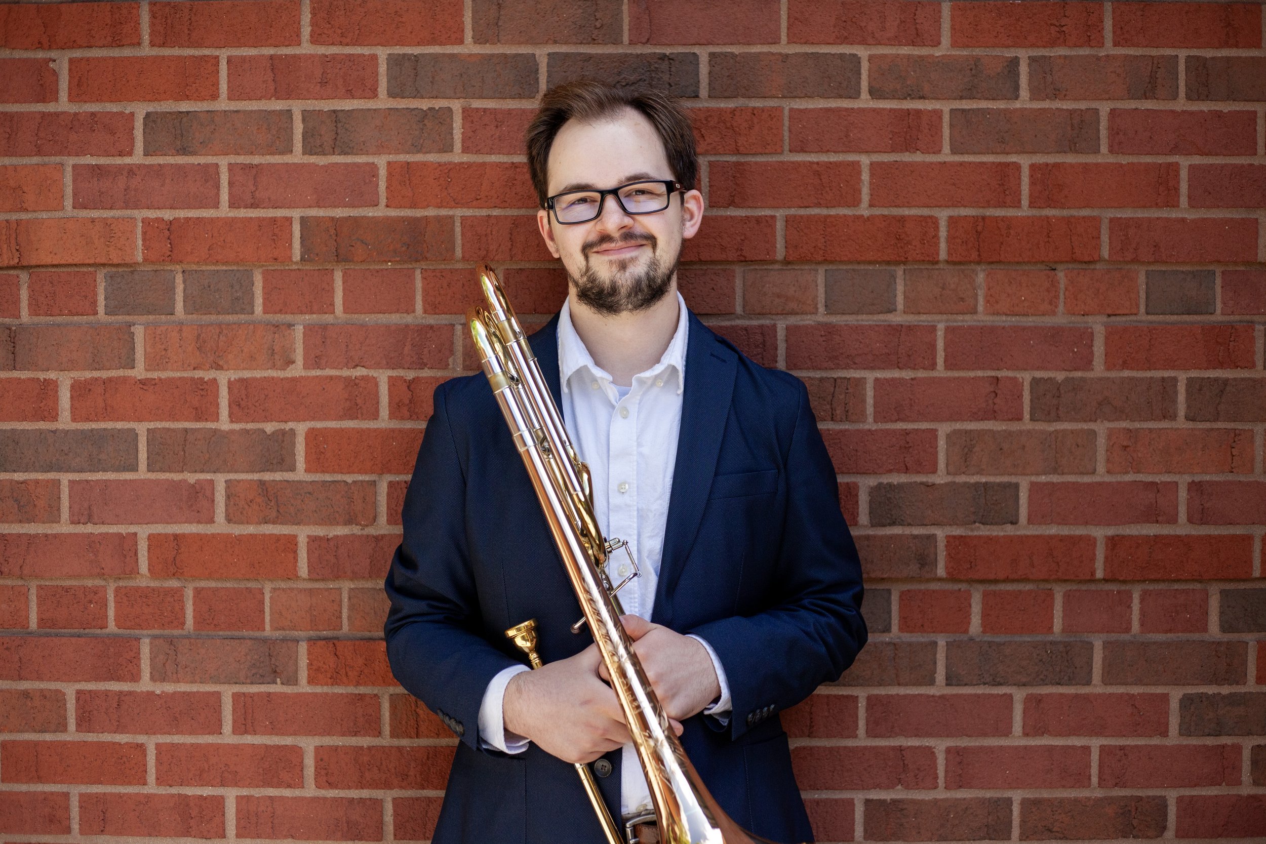 A man with glasses, a beard, and brown hair, dressed in a dark suit with a white shirt, holding a brass instrument, standing against a red brick wall.