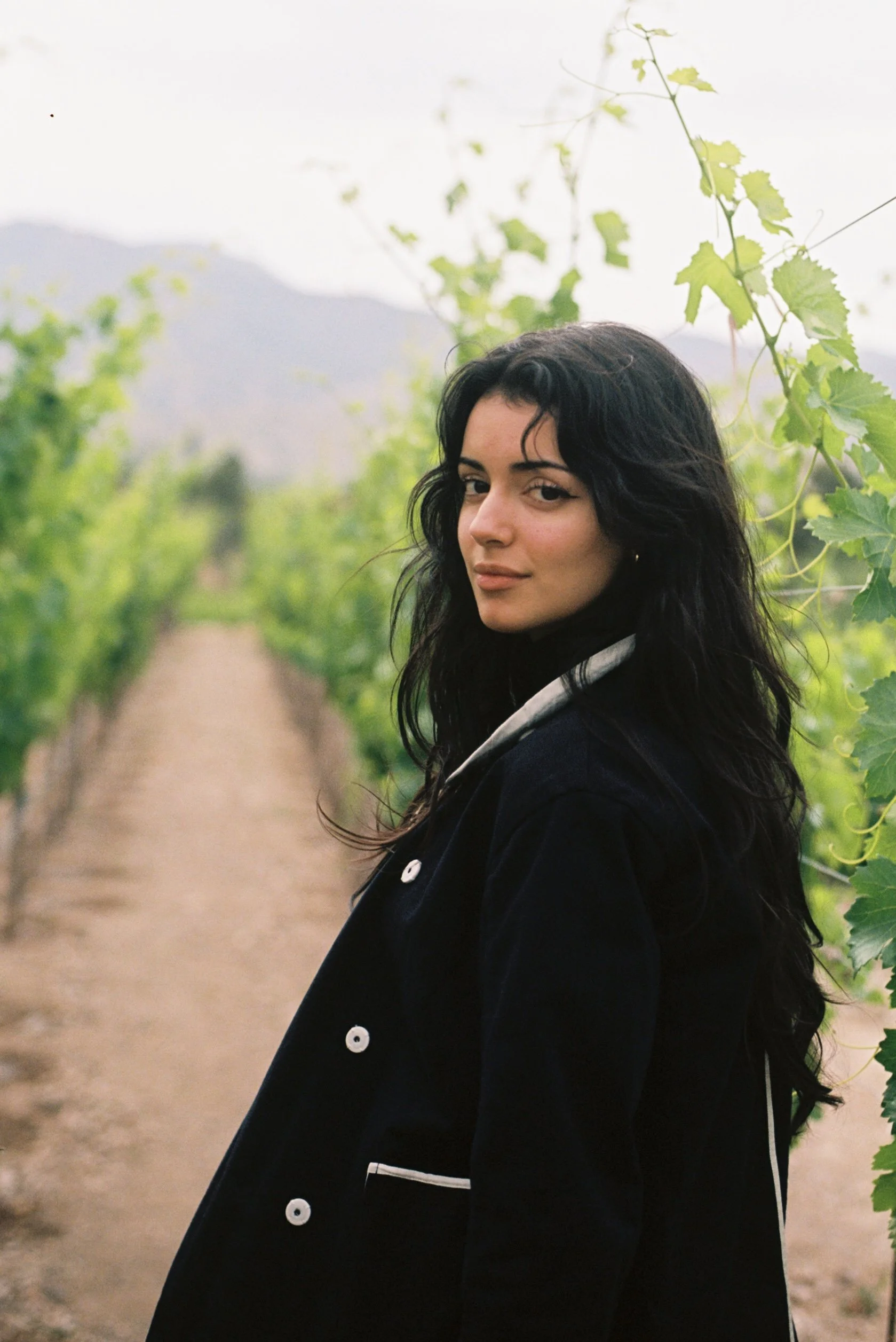 A young woman with long dark hair standing on a dirt path in a vineyard surrounded by green grapevines, with mountains in the background.
