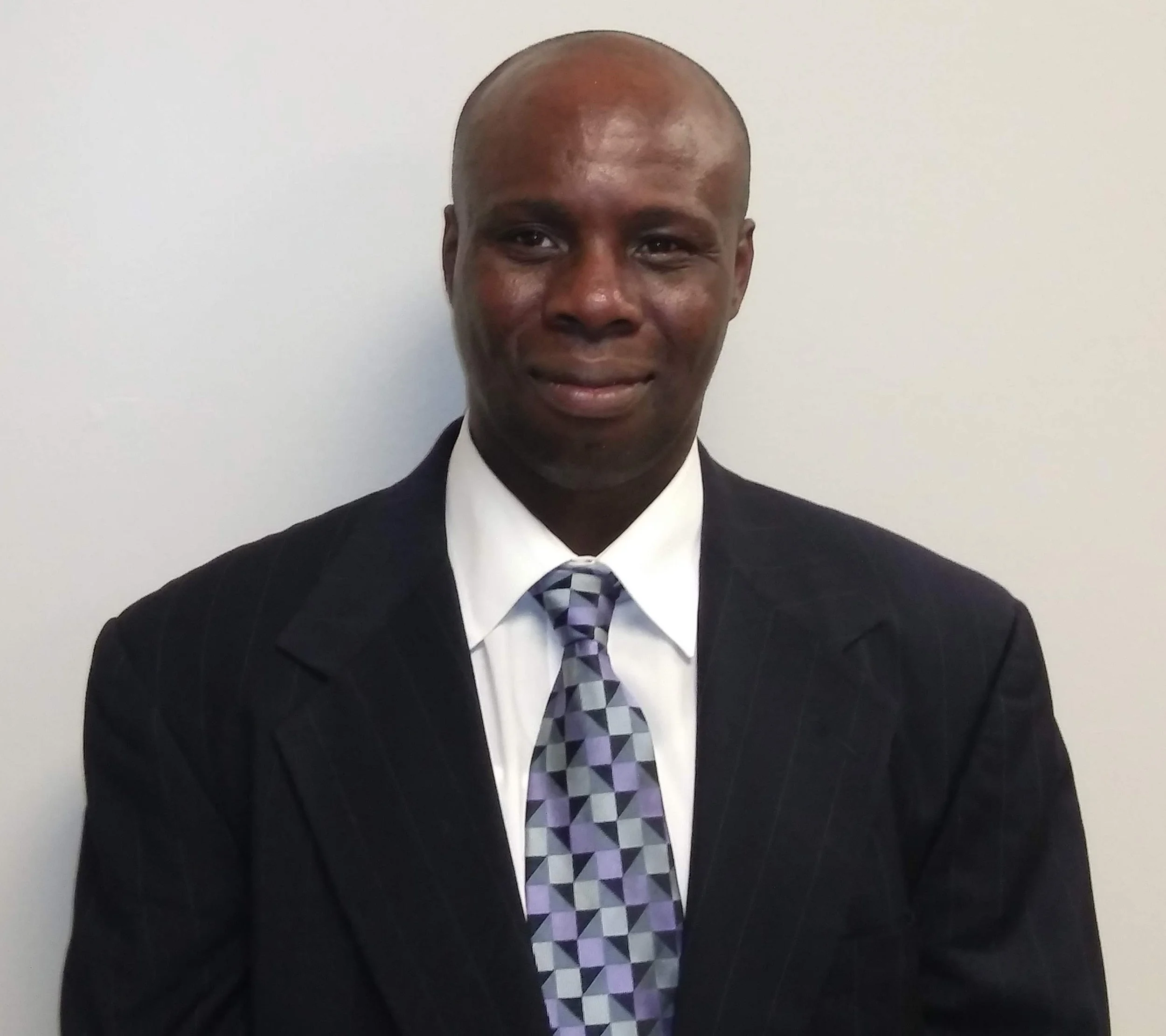 A man in a business suit, white shirt, and a patterned tie, standing against a plain white wall, smiling at the camera.