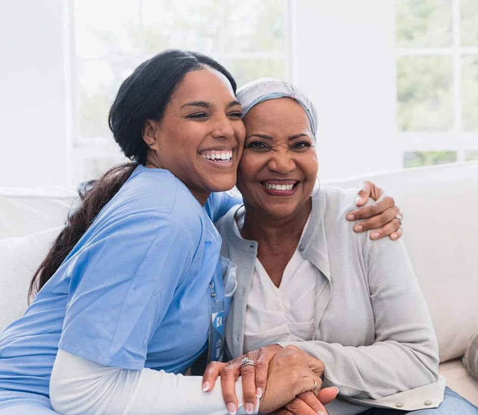 A young woman in scrubs hugging an older woman in a light-colored shirt and headscarf, both smiling warmly in a bright room.