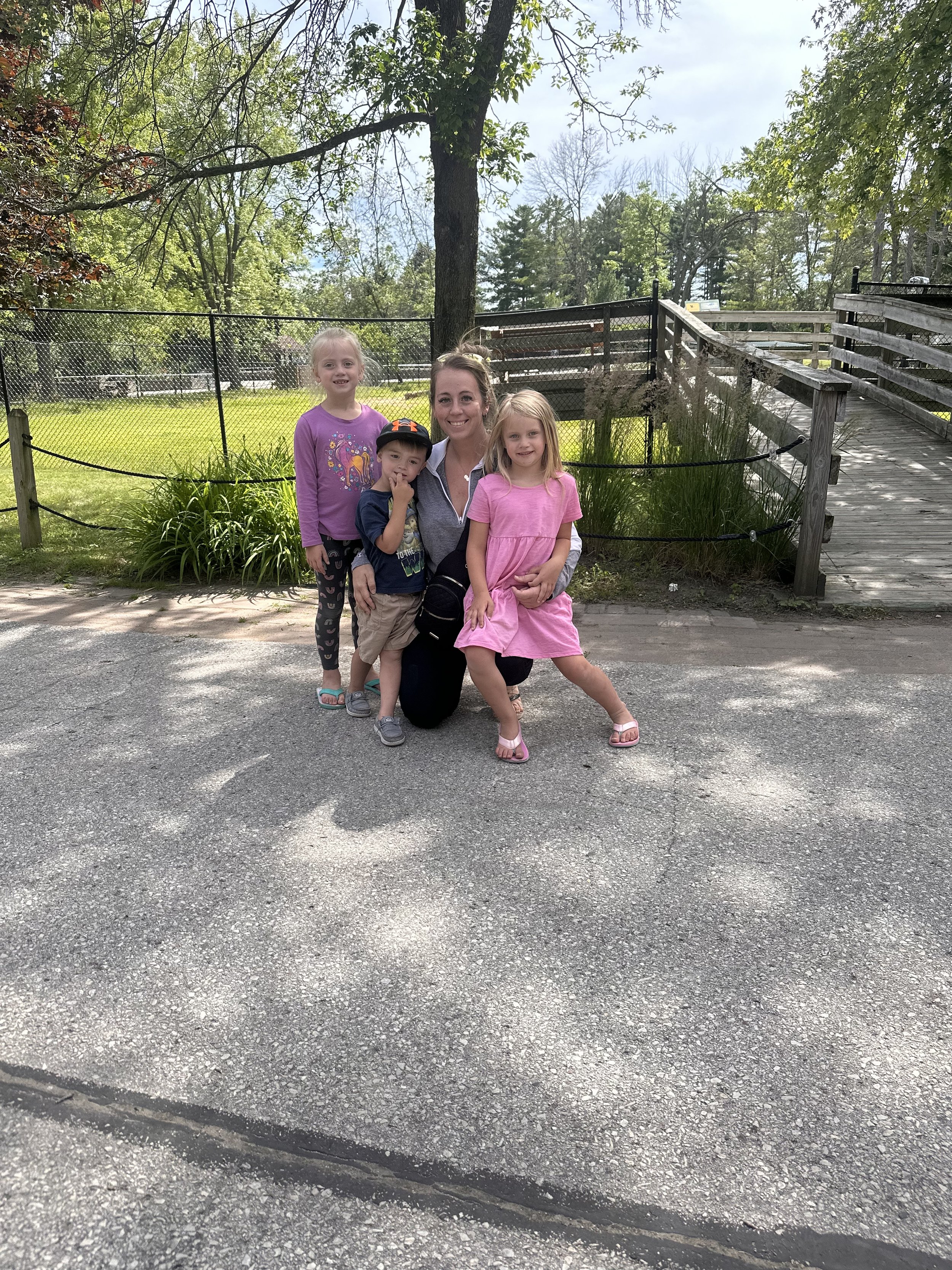 Children and a woman posing for a photo outdoors in a park-like setting with trees, grass, a wooden ramp, and a chain-link fence.