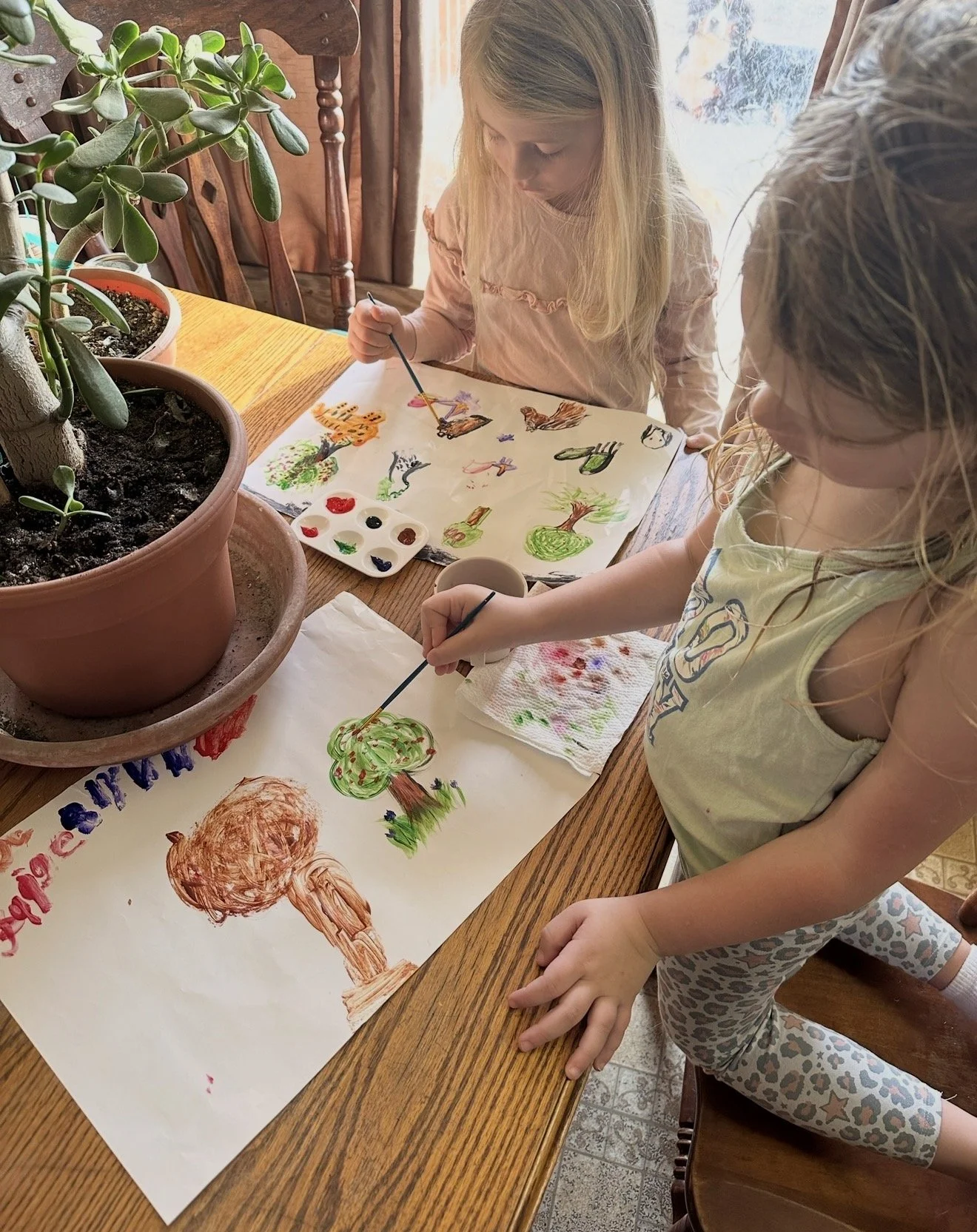 Two young girls are painting and drawing colorful pictures of trees, animals, and other objects on large sheets of white paper at a wooden table. There are potted plants on the table.