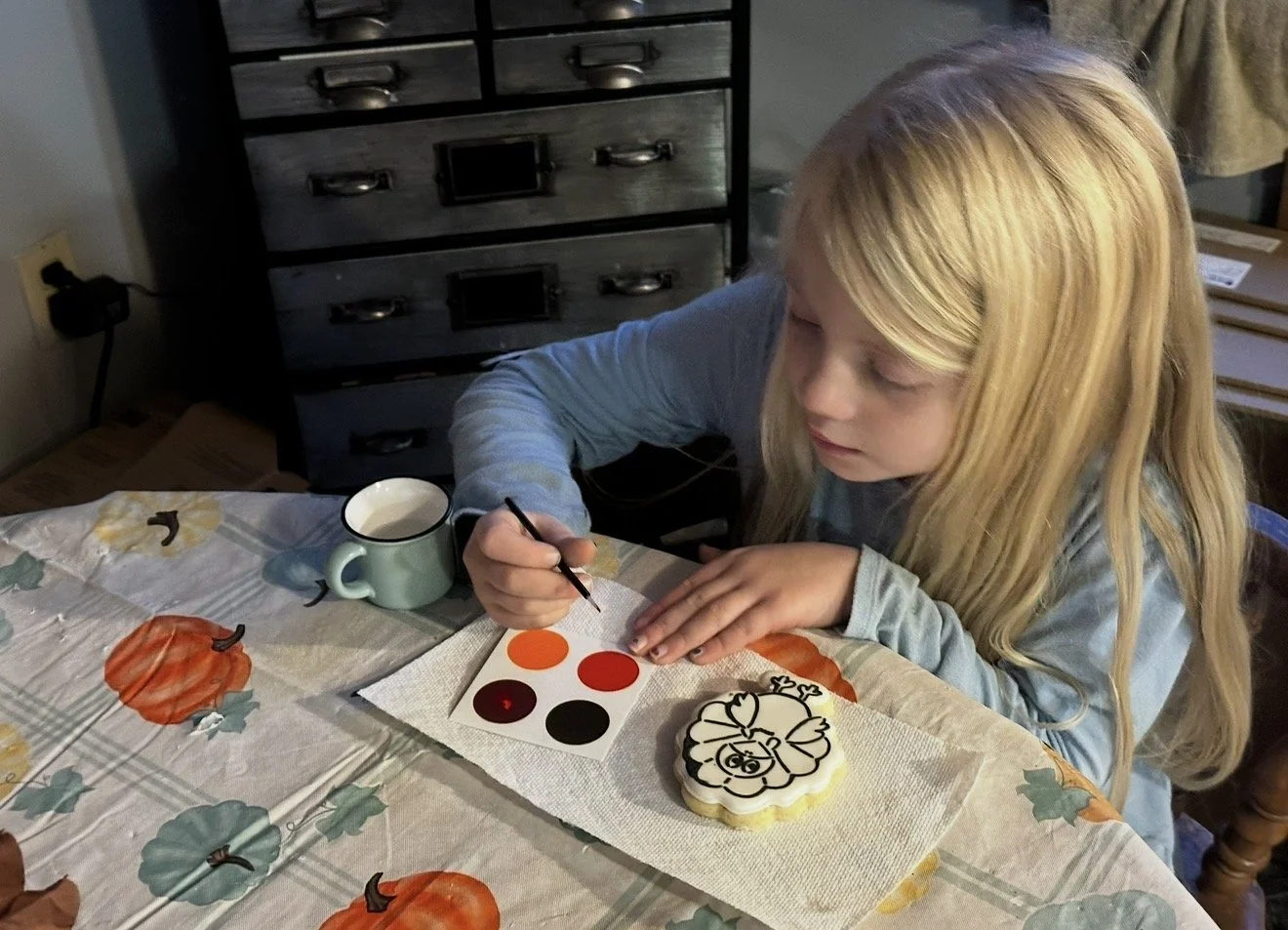 A young girl with long blonde hair, painting a pumpkin-shaped cookie with black icing. The table is decorated with a fall-themed tablecloth featuring pumpkins and leaves. There is a mug of water, a color palette, and a cookie with a pumpkin face drawing.