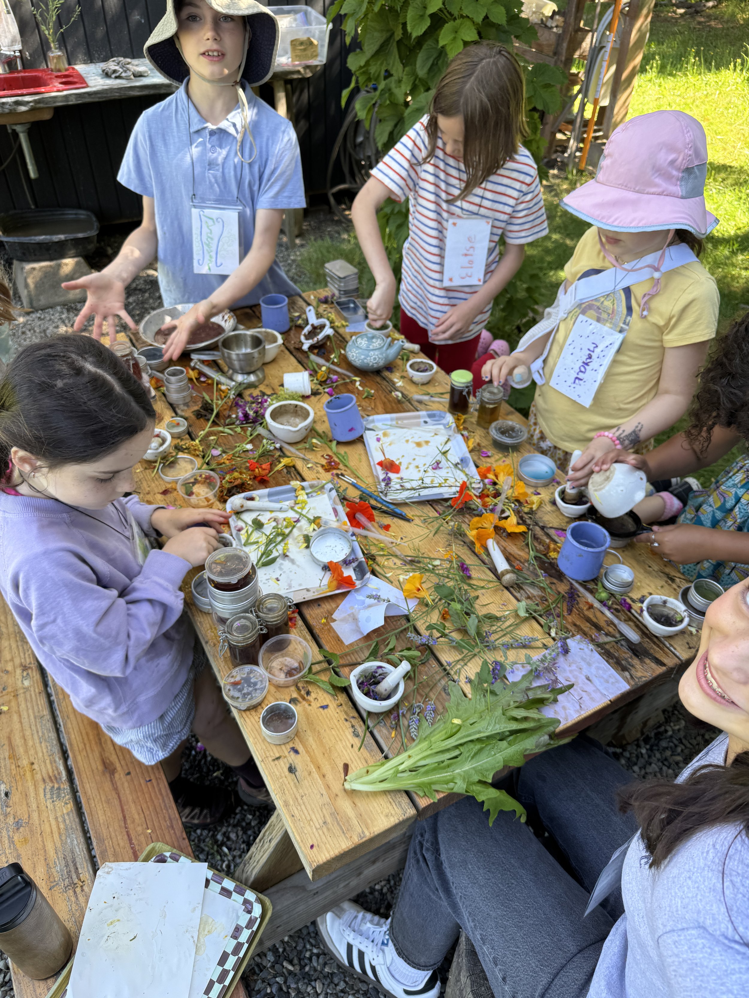 Group of children engaging in an outdoor flower arranging activity at a wooden table, decorated with flowers, jars, and gardening supplies, in a garden setting.