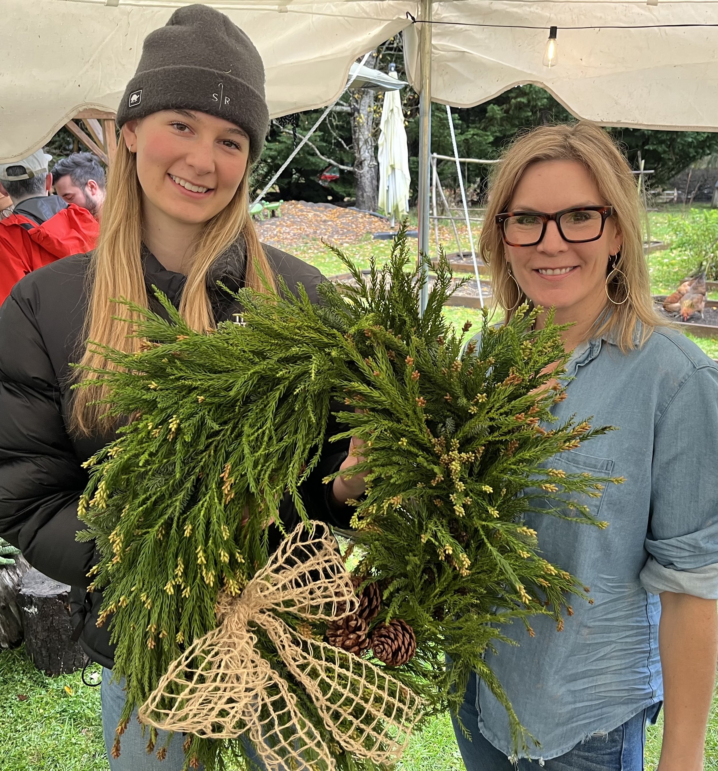 Two women holding a Christmas tree and smiling outdoors under a canopy with string lights. One woman wears a gray beanie and black jacket, the other has glasses and blonde hair.