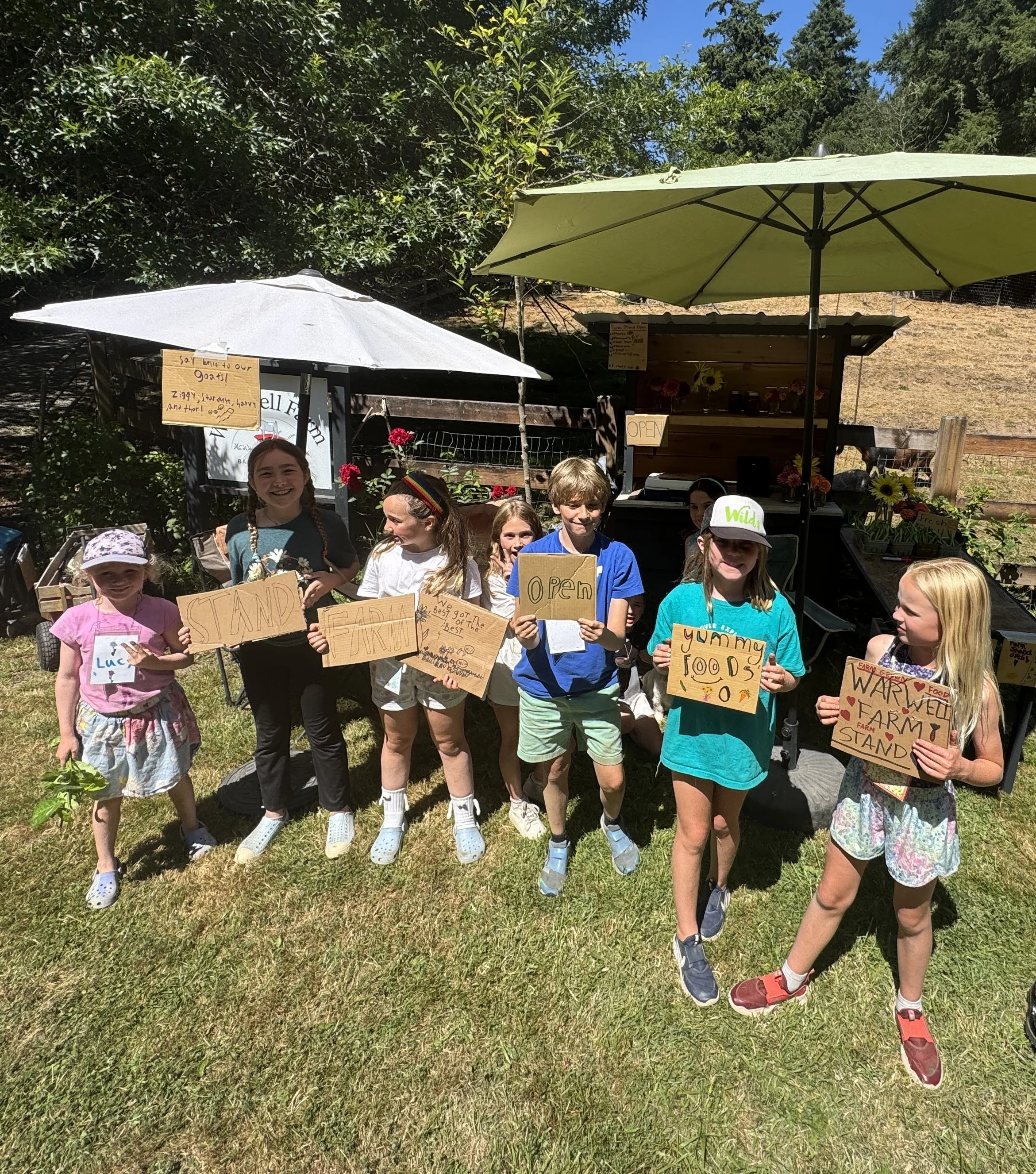Young children standing outdoors on grass, holding signs promoting farm stand and farm stand food, with trees, umbrellas, and a small farm stand in the background.
