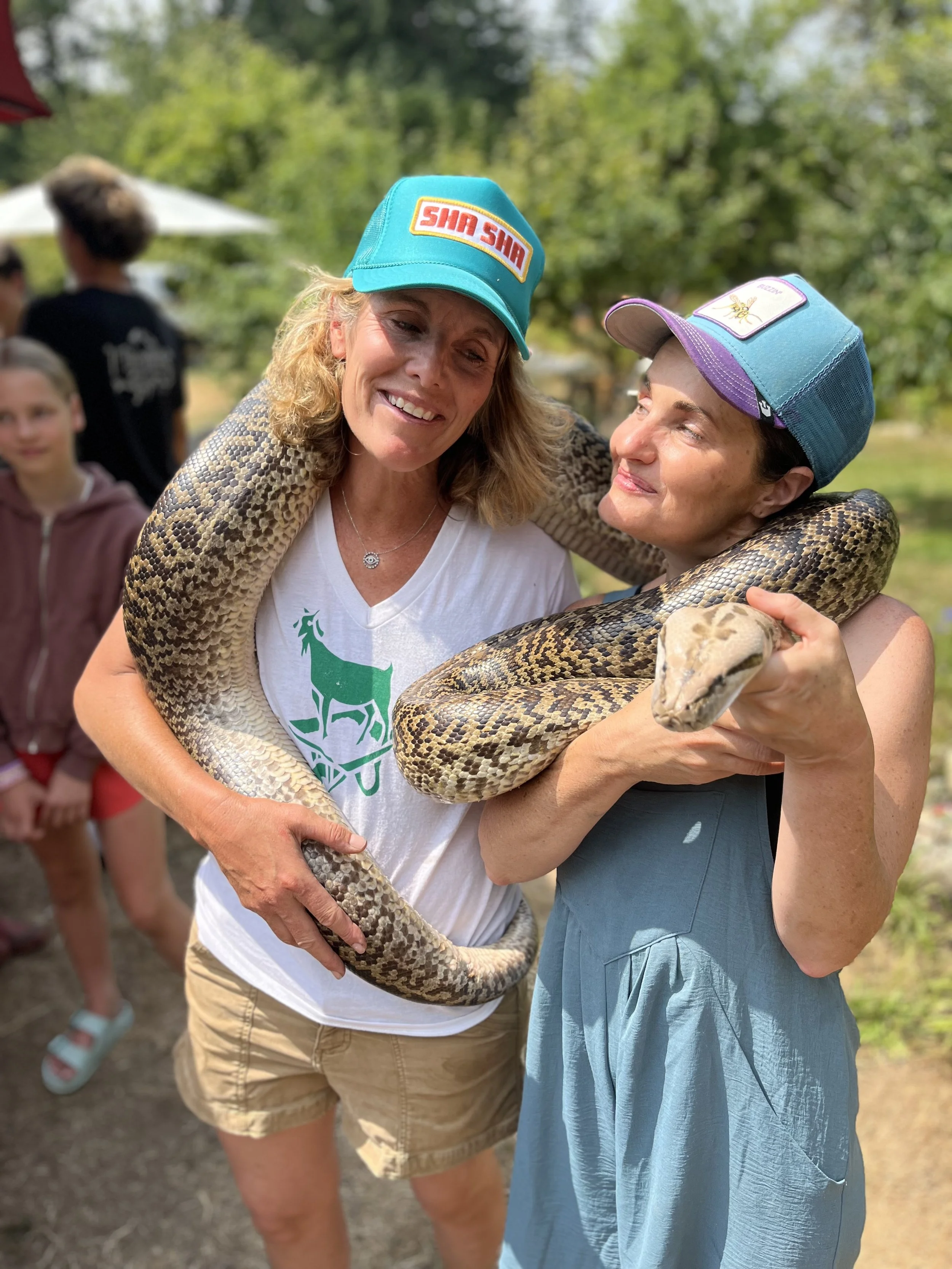 Two women smiling and hugging a large snake outdoors during daytime. One woman is wearing a blue cap with a bee patch, and the other a turquoise cap with a logo. In the background, there are people and greenery.