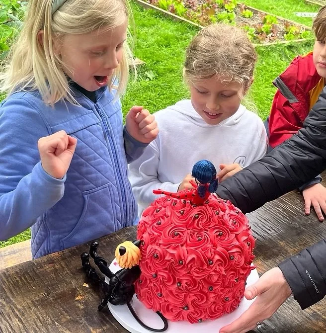 Three children gathered around a large, red, rosette-covered cake with a Barbie doll on top, celebrating outdoors with a wooden table and green grass in the background.