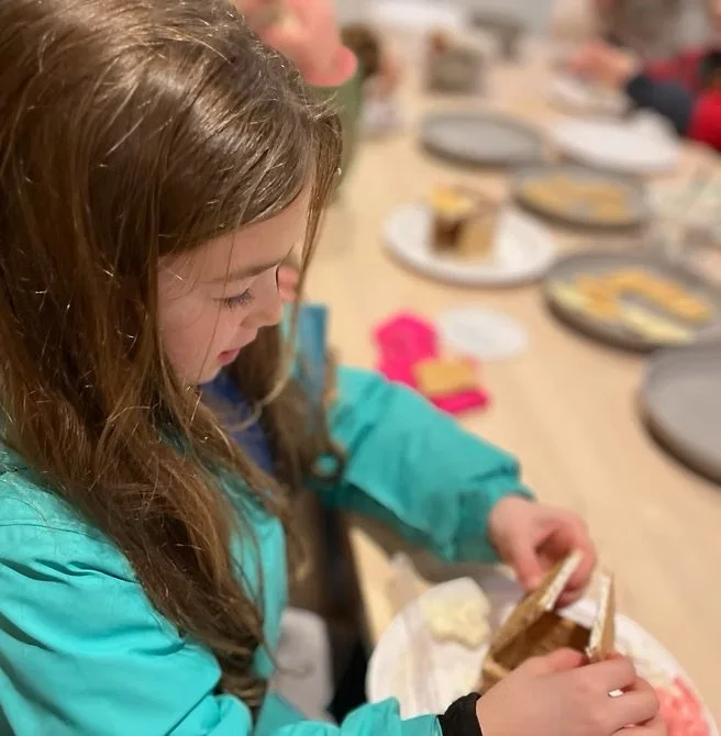 A young girl in a teal jacket opening a package of graham crackers at a table set with plates, desserts, and pink napkins.