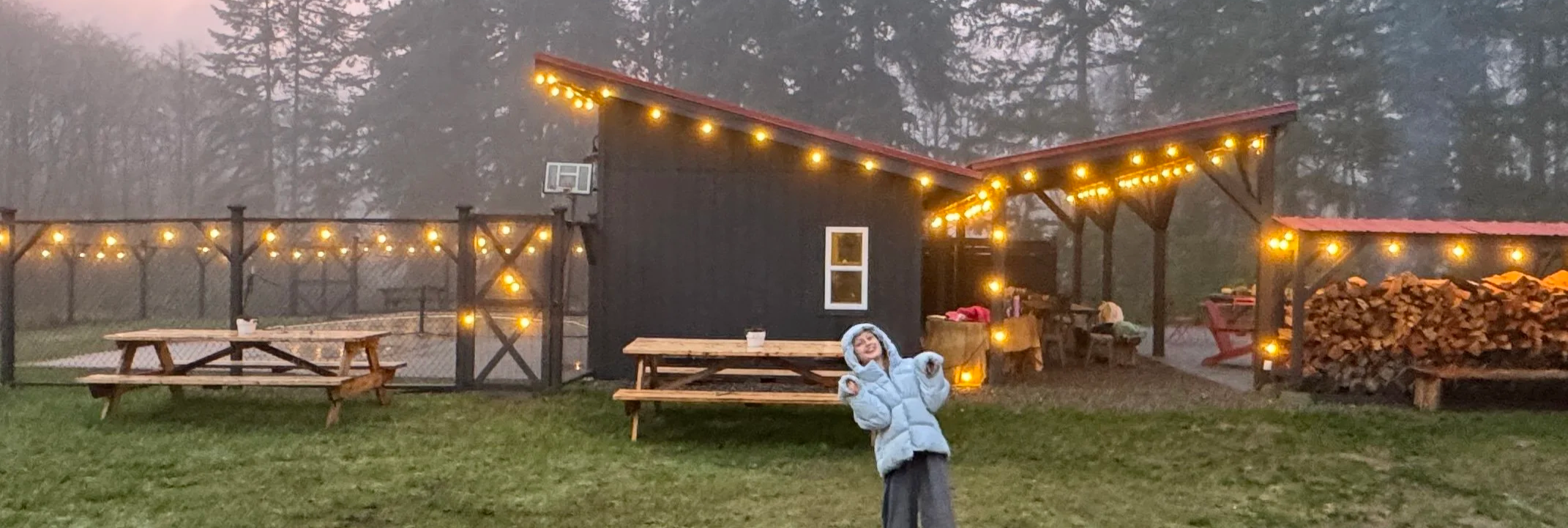 A child in a winter coat smiling and making playful gestures in front of a backyard with string lights, picnic tables, and a black building with a red roof at dusk or dawn.