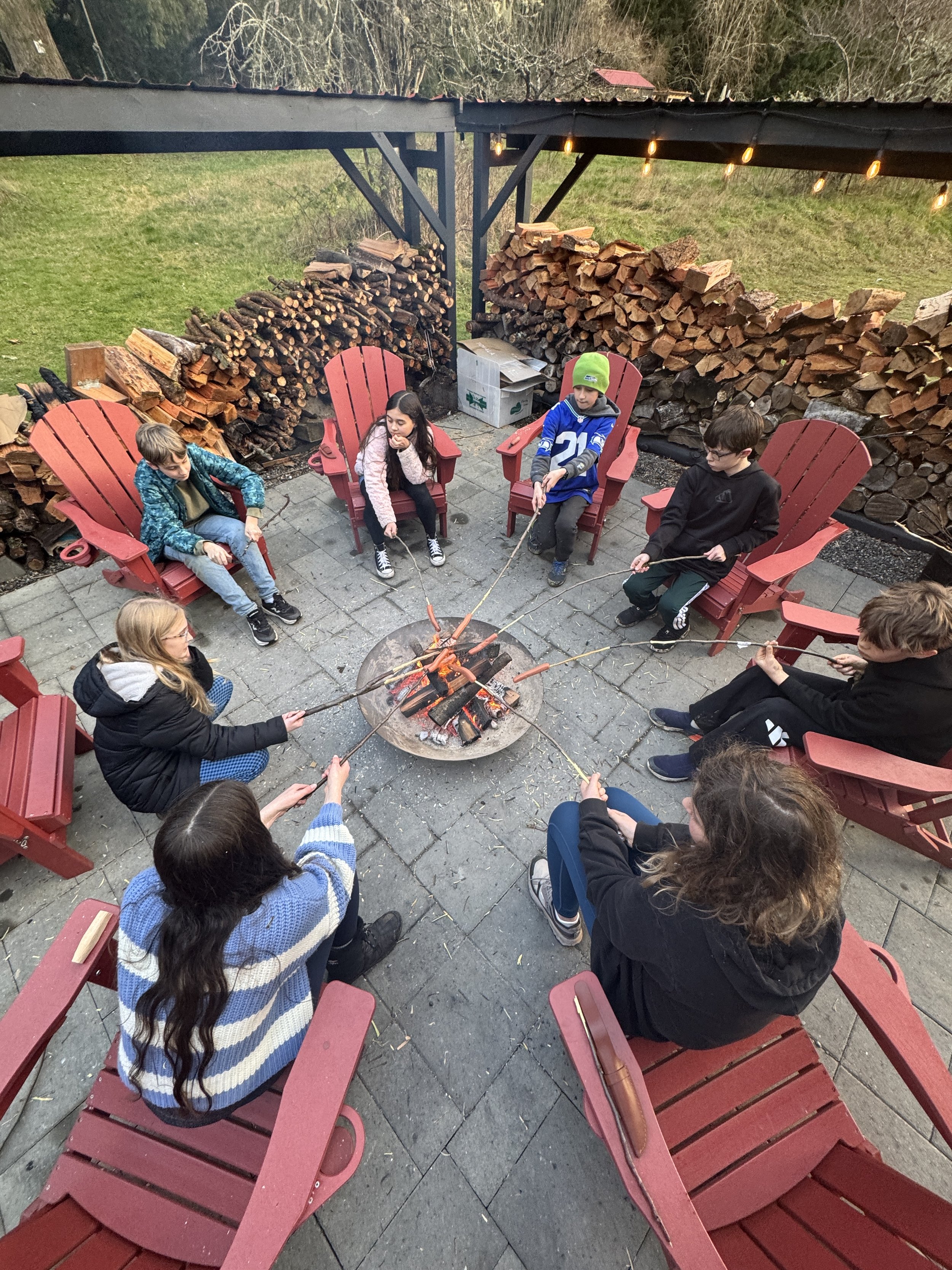 A group of children sitting in red Adirondack chairs around a fire pit roasting marshmallows on sticks. Wood logs are stacked against a black wooden structure with hanging lights, and a grassy hill with trees is visible in the background.