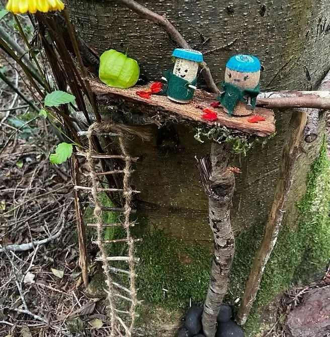 Miniature craft scene on tree trunk with two small figurines wearing blue hats, a green leaf, some red flower petals, a small green object, and a makeshift ladder leaning against the tree.