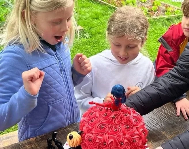 Three children gathered around a table with a red, rosette-patterned cake decorated with a Spider-Man figurine topper. The children look excited and happy.