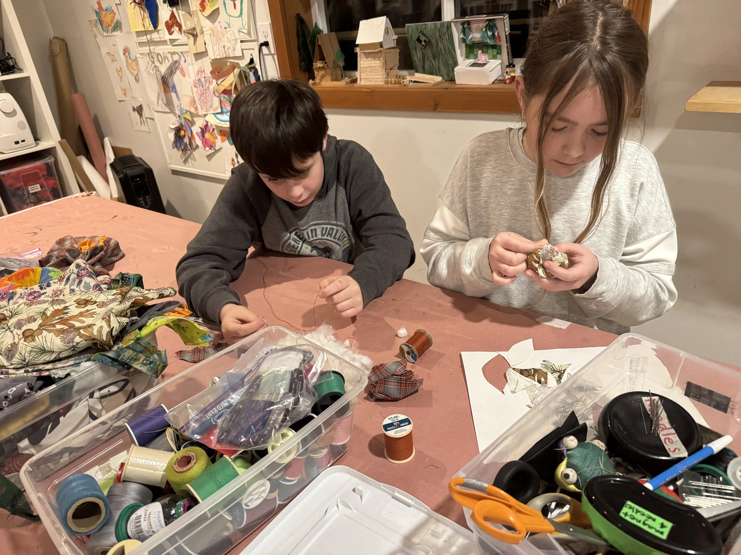 Children sitting at a table engaging in arts and crafts, surrounded by craft supplies such as thread, fabric, scissors, and tape.