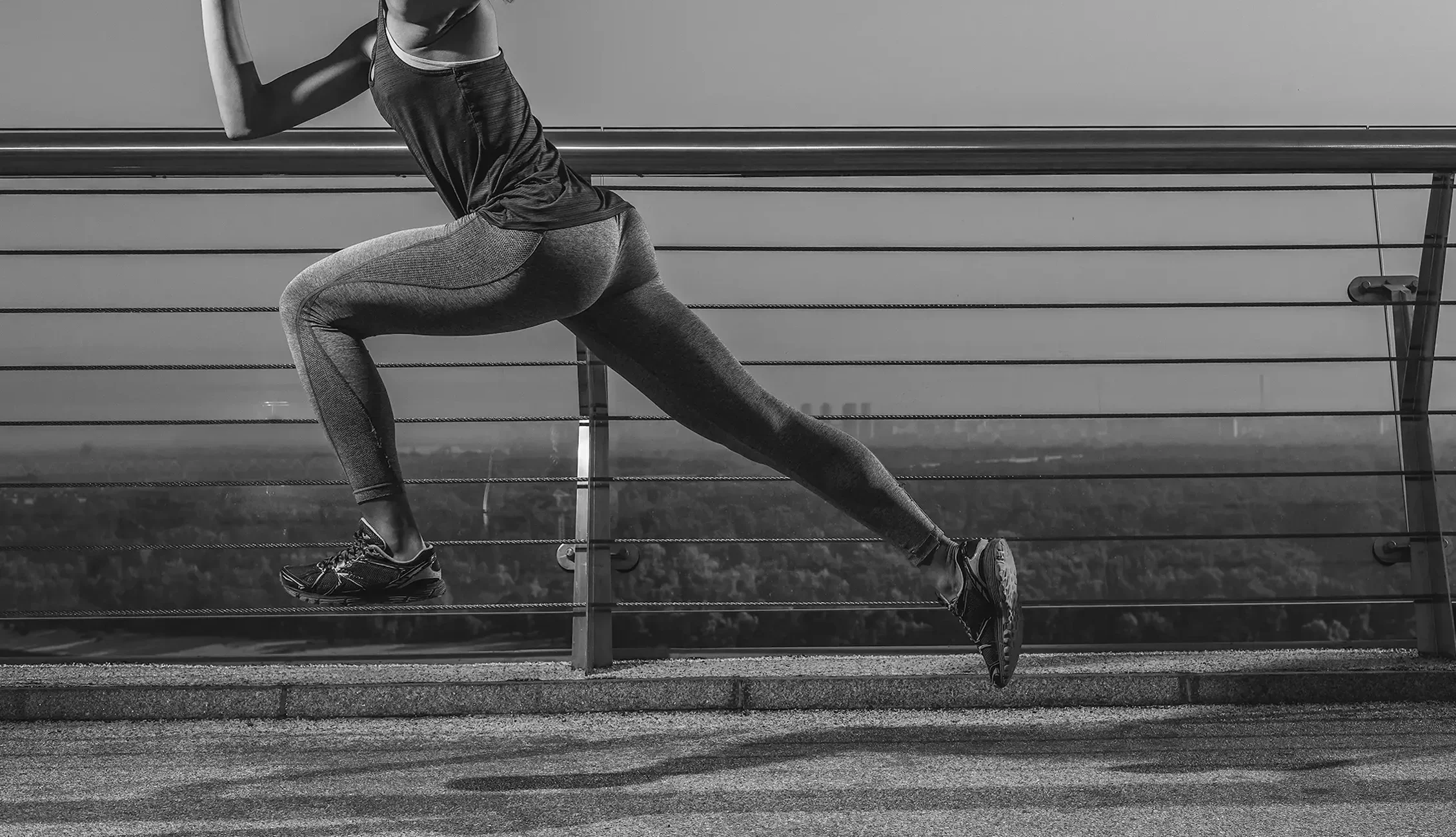A person wearing athletic clothing and running shoes lunges on a track in front of a metal guardrail with a scenic view in the background.