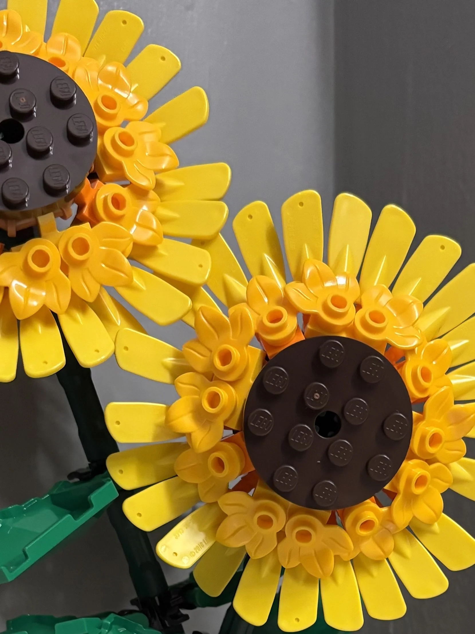 Close-up of a flower-shaped structure made from yellow, orange, green, and black plastic LEGO bricks, arranged to resemble a sunflower.