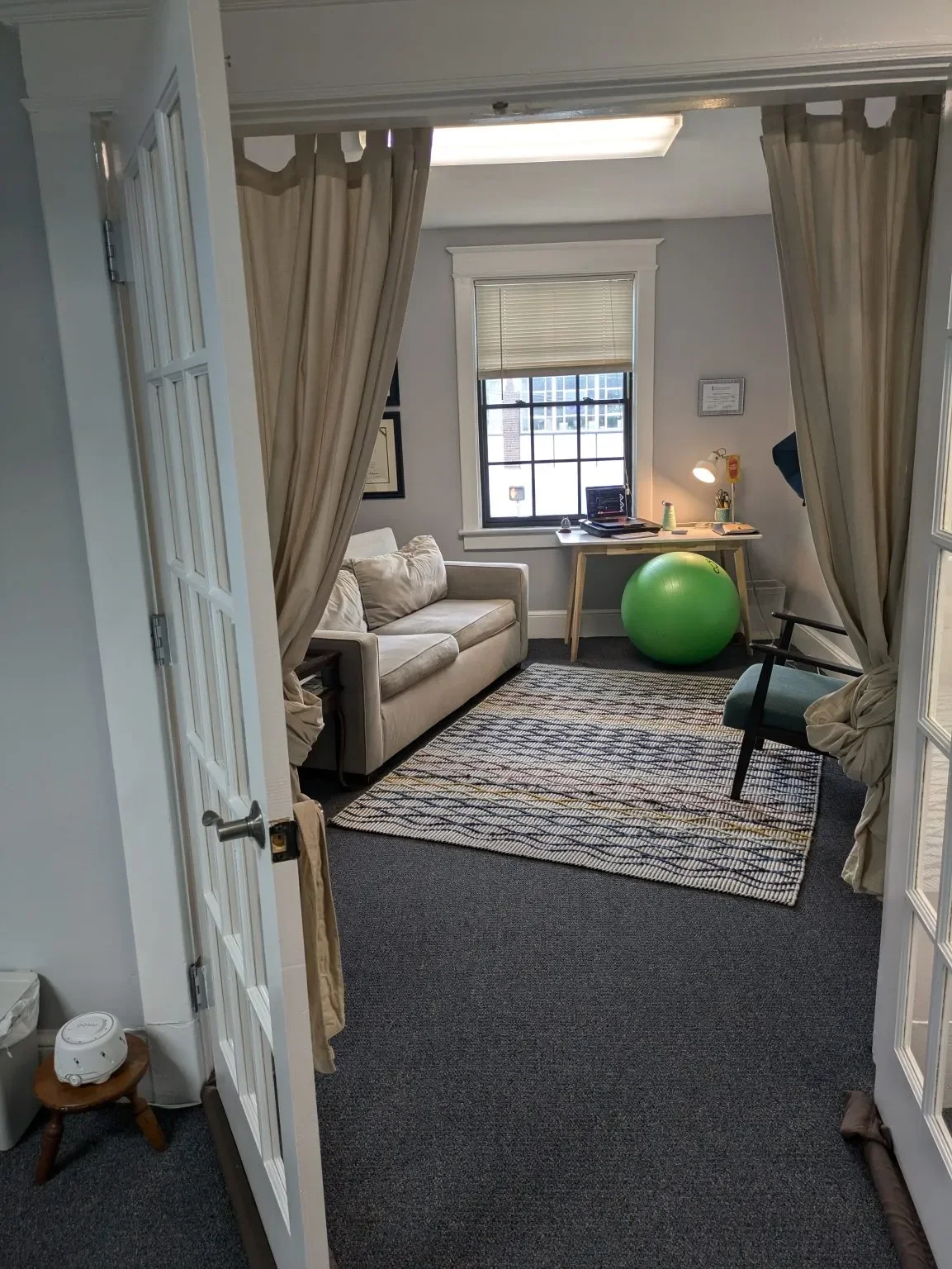 Shows the entrance to the therapy space, with double doors, a beige couch, green exercise ball and basic desk, colorful rug and additional seating.