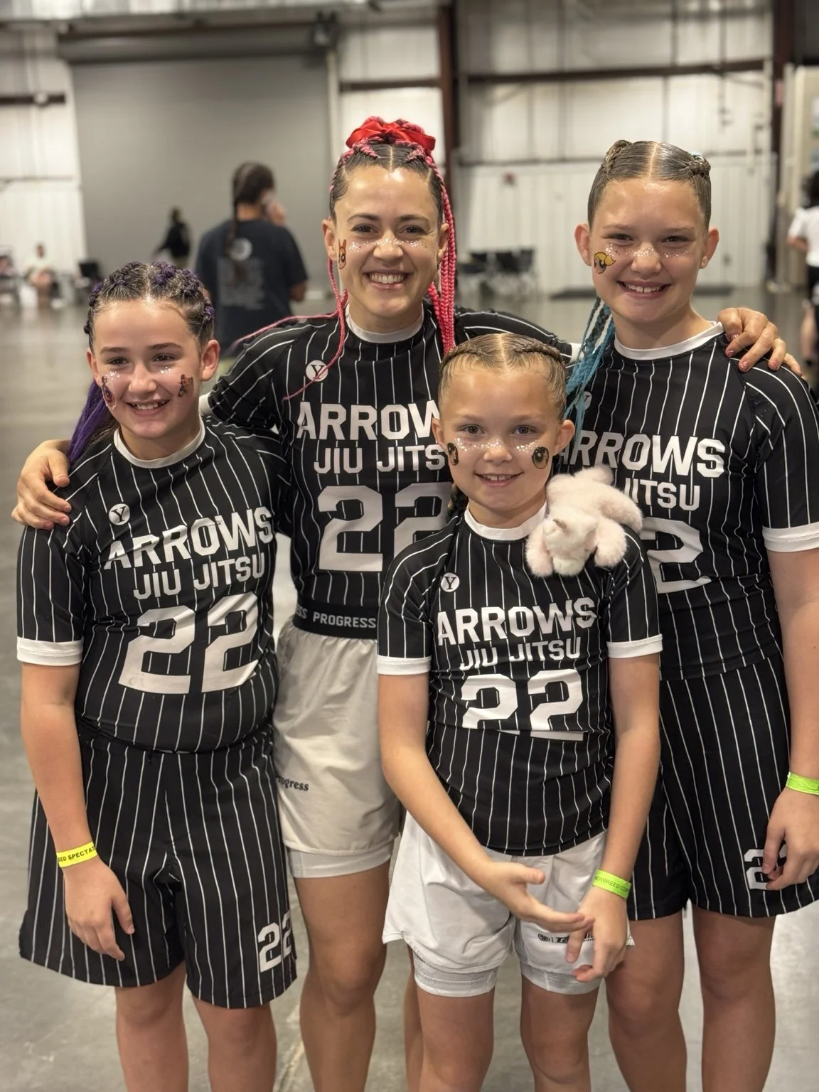Group of five children and a woman in black and white striped sports outfits inside a gym or sports hall. The children and woman are smiling, with some face paint and one child holding a stuffed animal. They are part of a team called 'Arrows Jiu Jitsu'.
