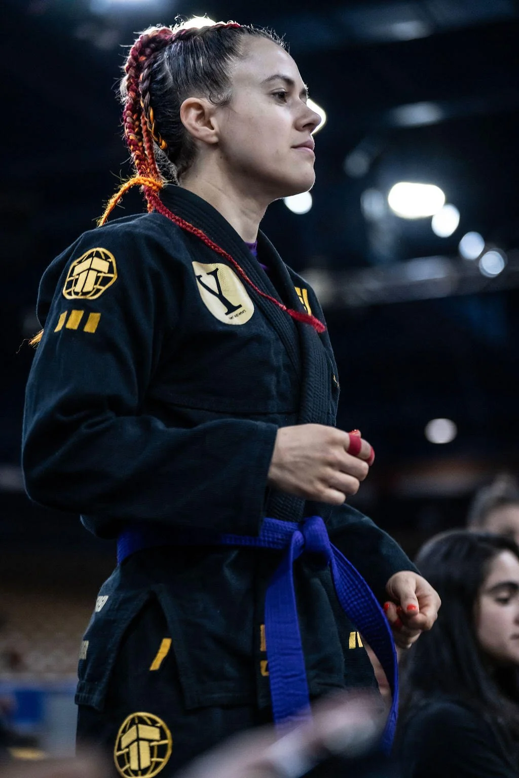 A woman with braided hair in a Brazilian Jiu-Jitsu gi with patches, standing in a gym or competition setting with bright lights.