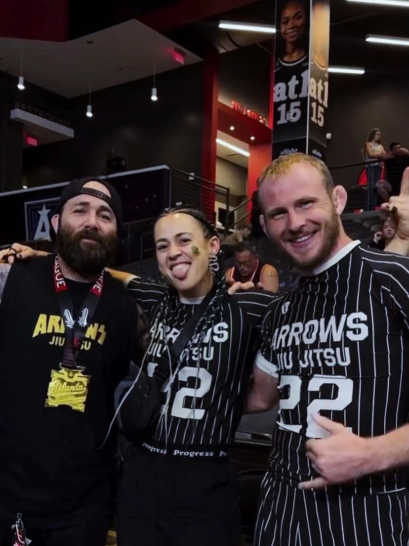 Three people at an indoor event, wearing black and white striped jerseys with 'ARROWS JIU JITSU' and numbers 12 and 22, smiling and posing for the camera. One person has a sticker on his face, another is sticking out her tongue.