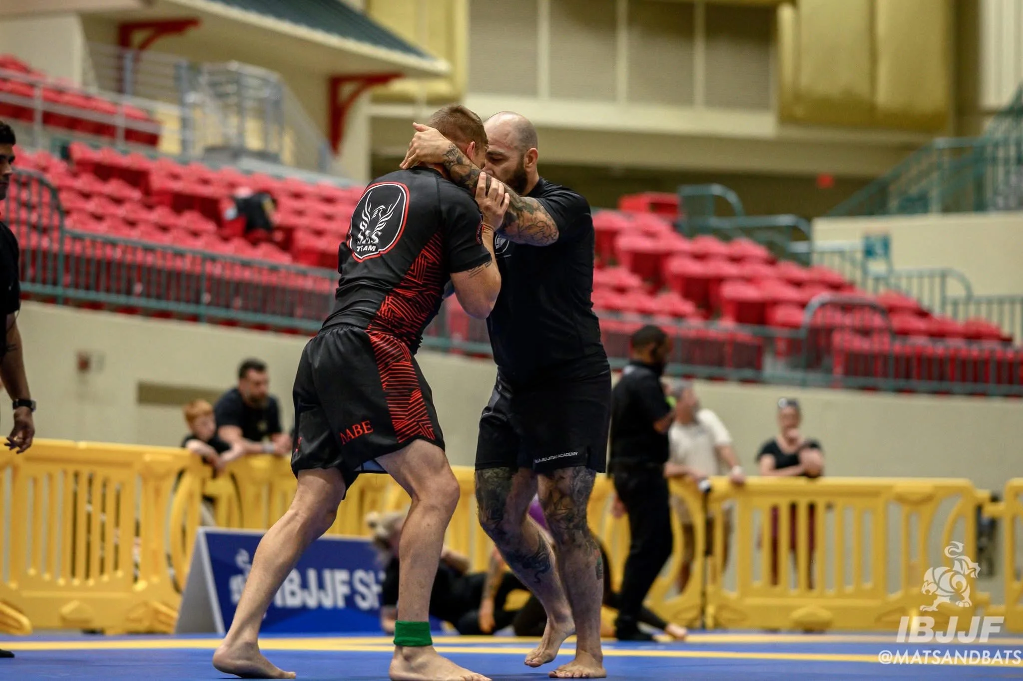 Two male mixed martial arts fighters grappling during a match in a gym or arena, with several spectators and officials in the background.