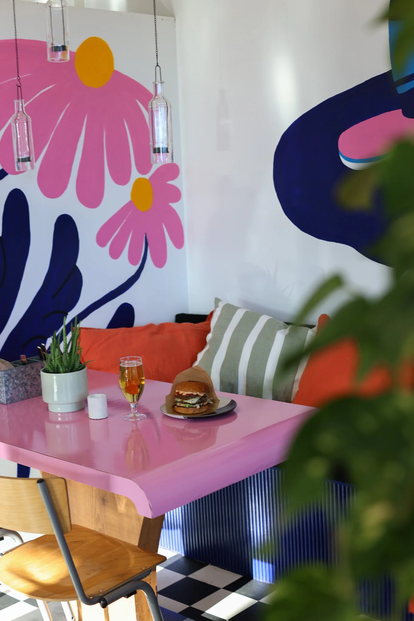 Värikäs ravintolasali, jossa pinkki pöytä, burgeri ja juomia, kasvi, seinämuraali, sohva ja tuoli.