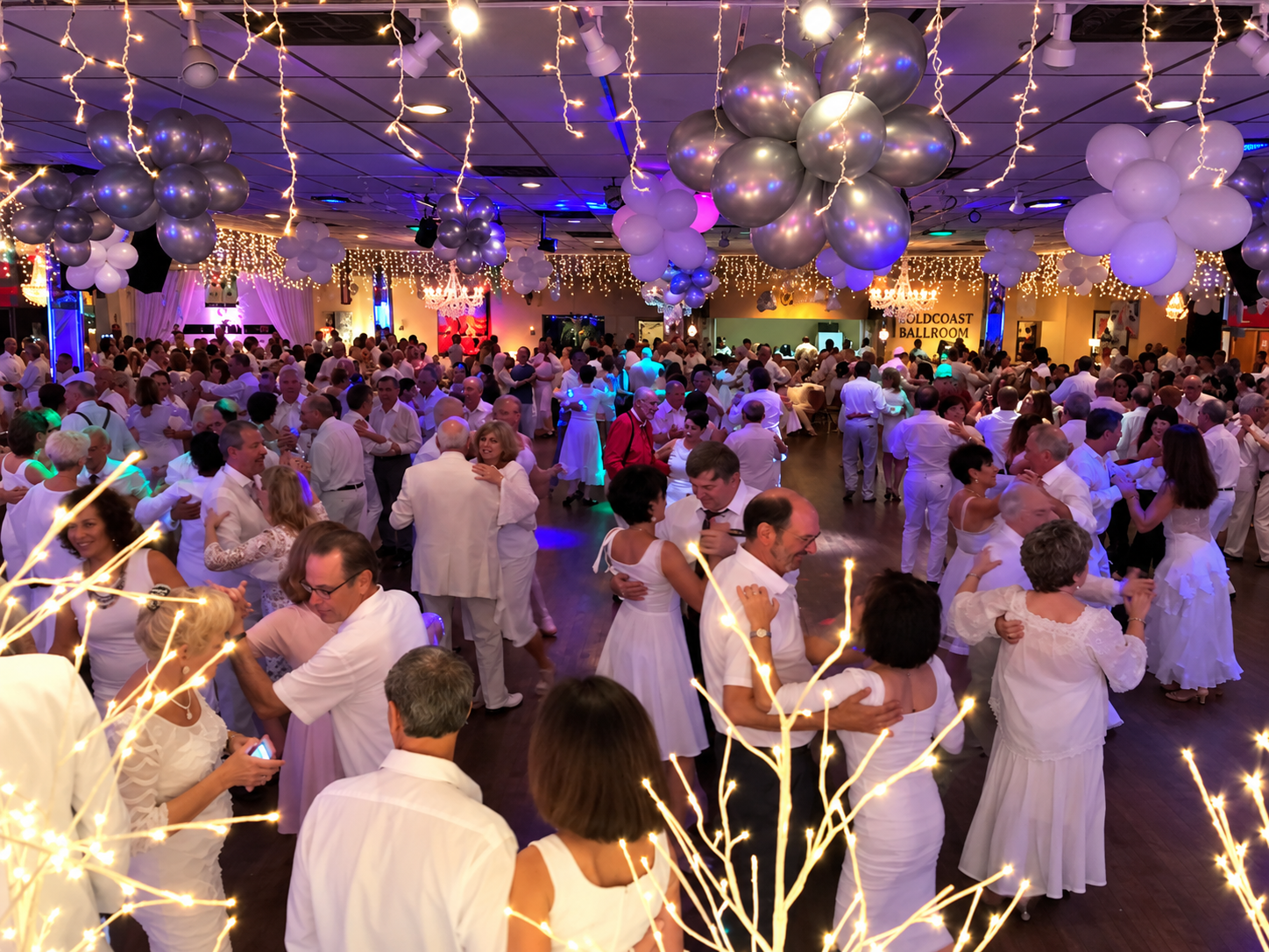 People dancing at a wedding reception in a decorated ballroom with balloons and string lights.