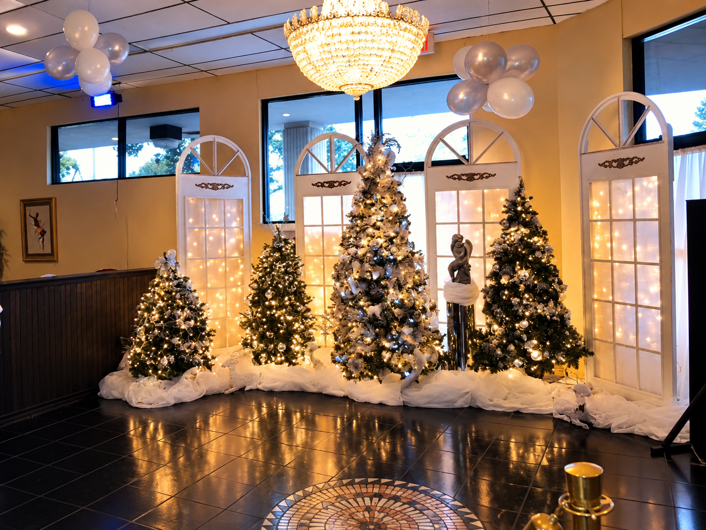 Indoor Christmas display with five decorated Christmas trees illuminated with white lights, set against a backdrop of white window-frames with fairy lights inside, and gold and white balloons hanging from the ceiling. There is a large chandelier overhead and a gold candle in the lower right corner.