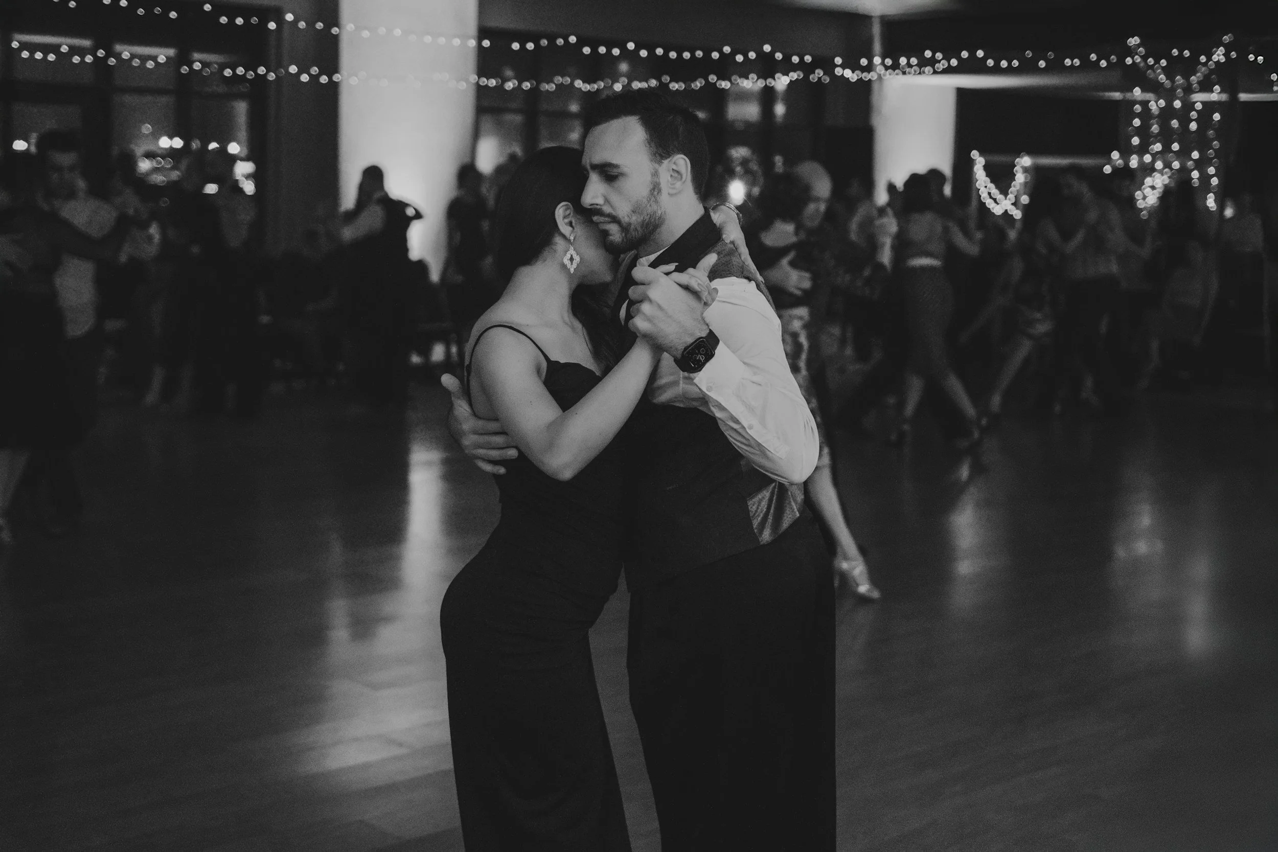 A man and woman dancing closely at a dimly lit party or dance hall decorated with string lights. The woman has long hair and is wearing earrings, while the man has facial hair and a watch.