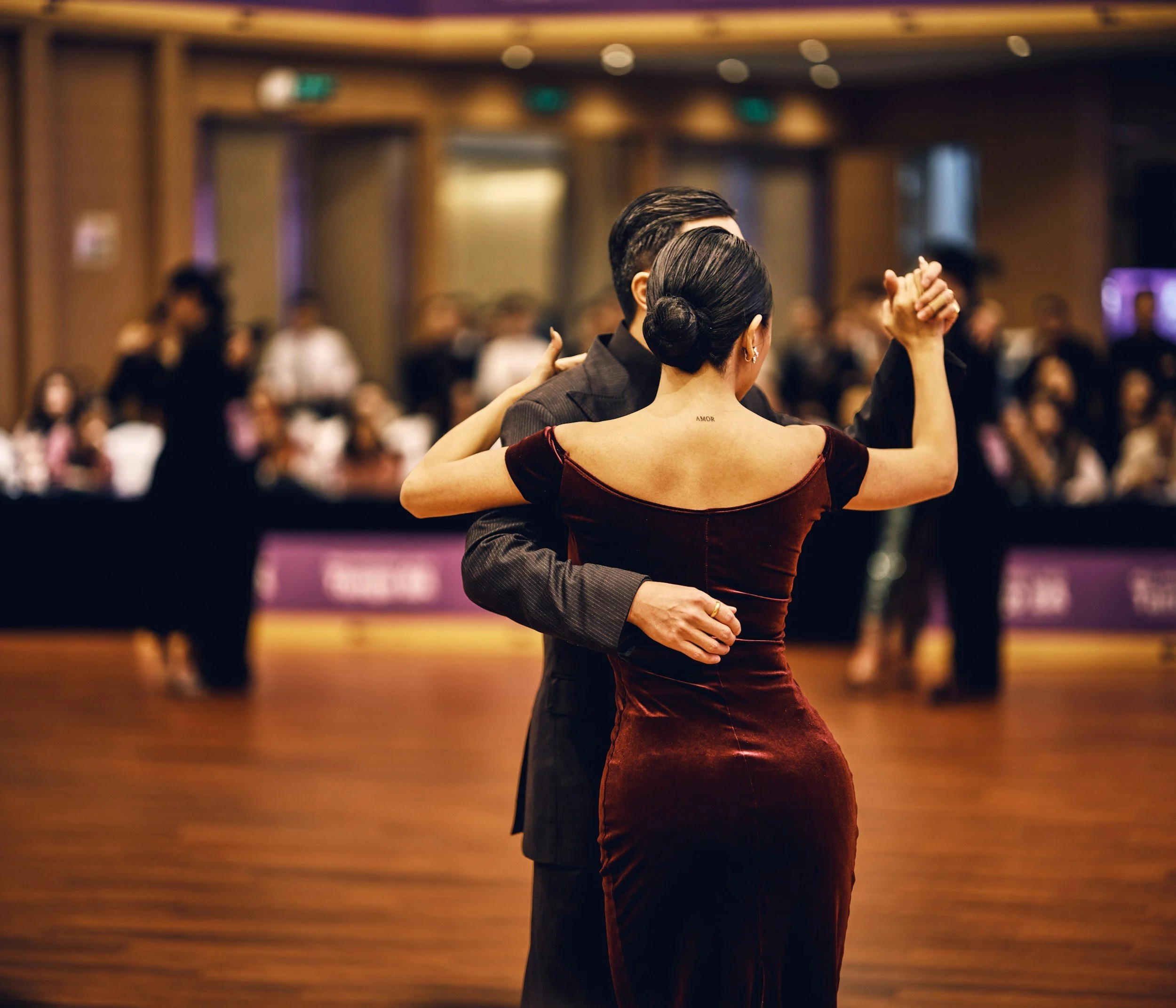 A couple dancing at a formal event in a ballroom, with an audience watching in the background.