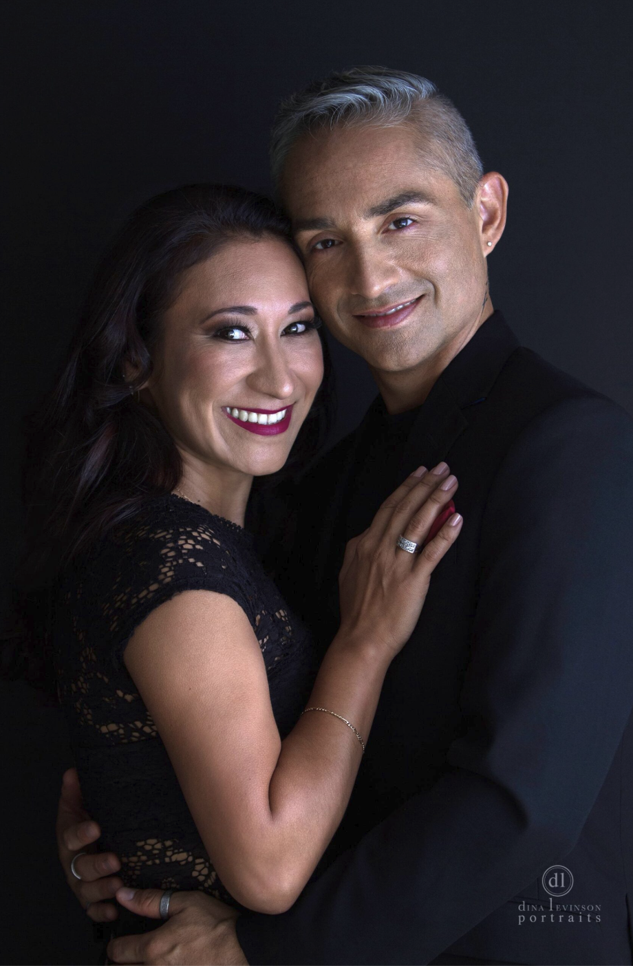 A smiling woman with dark hair and a woman with short, gray hair, dressed in black, embracing against a dark background.