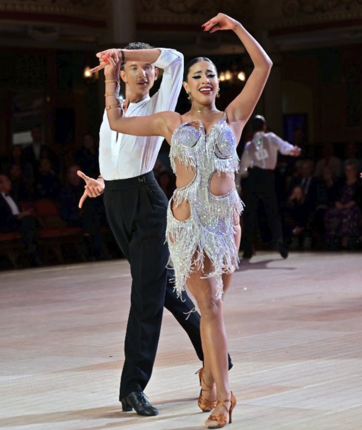 A couple performing a ballroom dance, with the woman in a sparkly, fringe dress and the man in a white shirt and black pants.