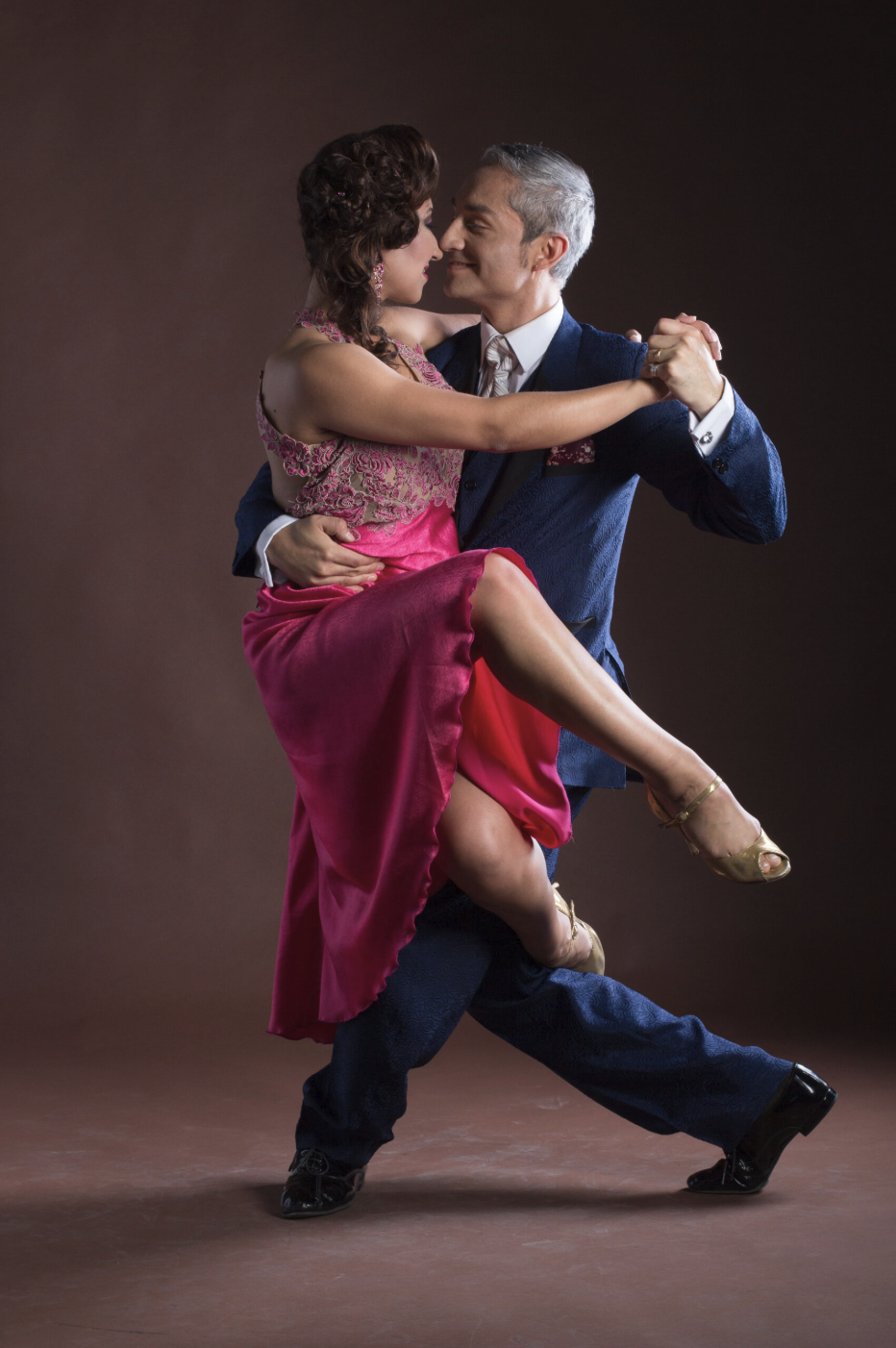 A man and a woman dance closely together, with the woman sitting on the man's lap, both smiling and looking at each other against a plain dark background.