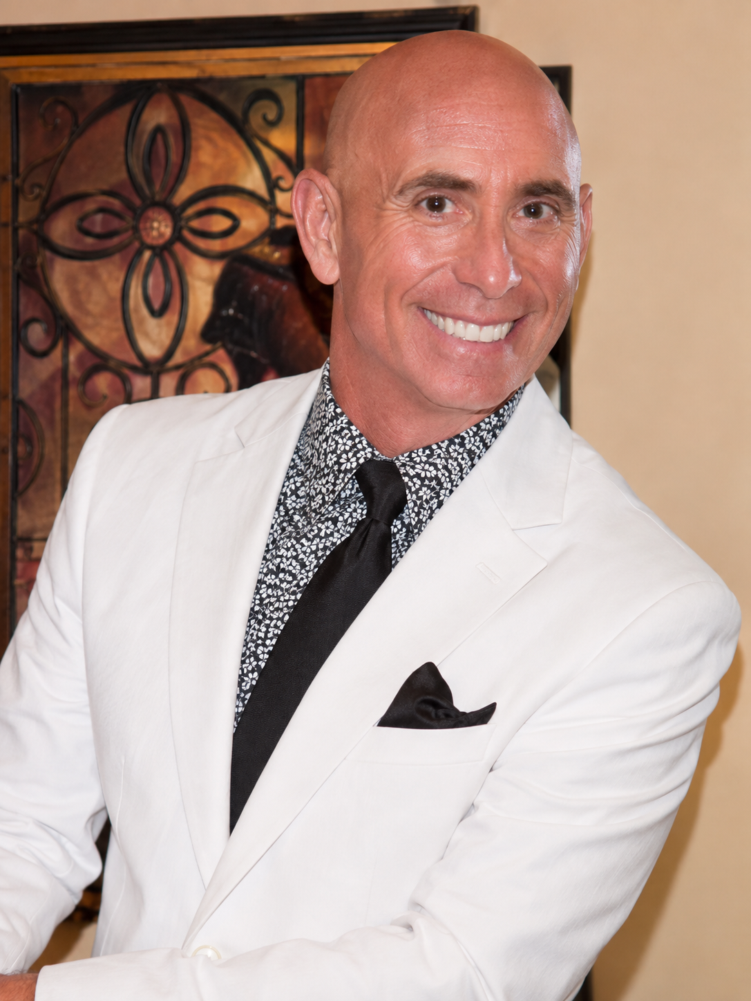 A man in a white suit with a black tie and patterned shirt, smiling, with artwork featuring a dog and a floral design in the background.