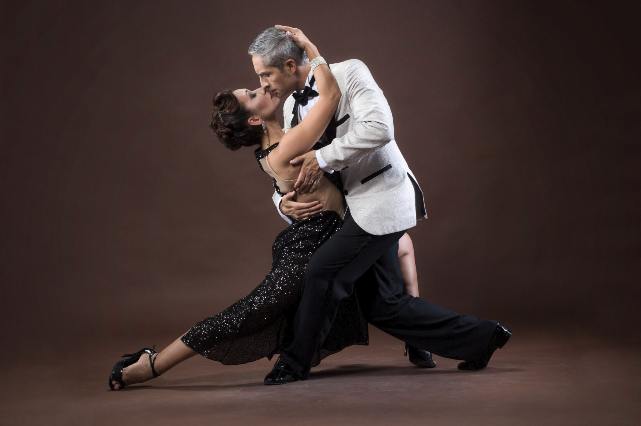 A man and woman dance closely, with the woman in a black sequined dress and heels, and the man in a light-colored tuxedo with a black bow tie, against a plain dark background.