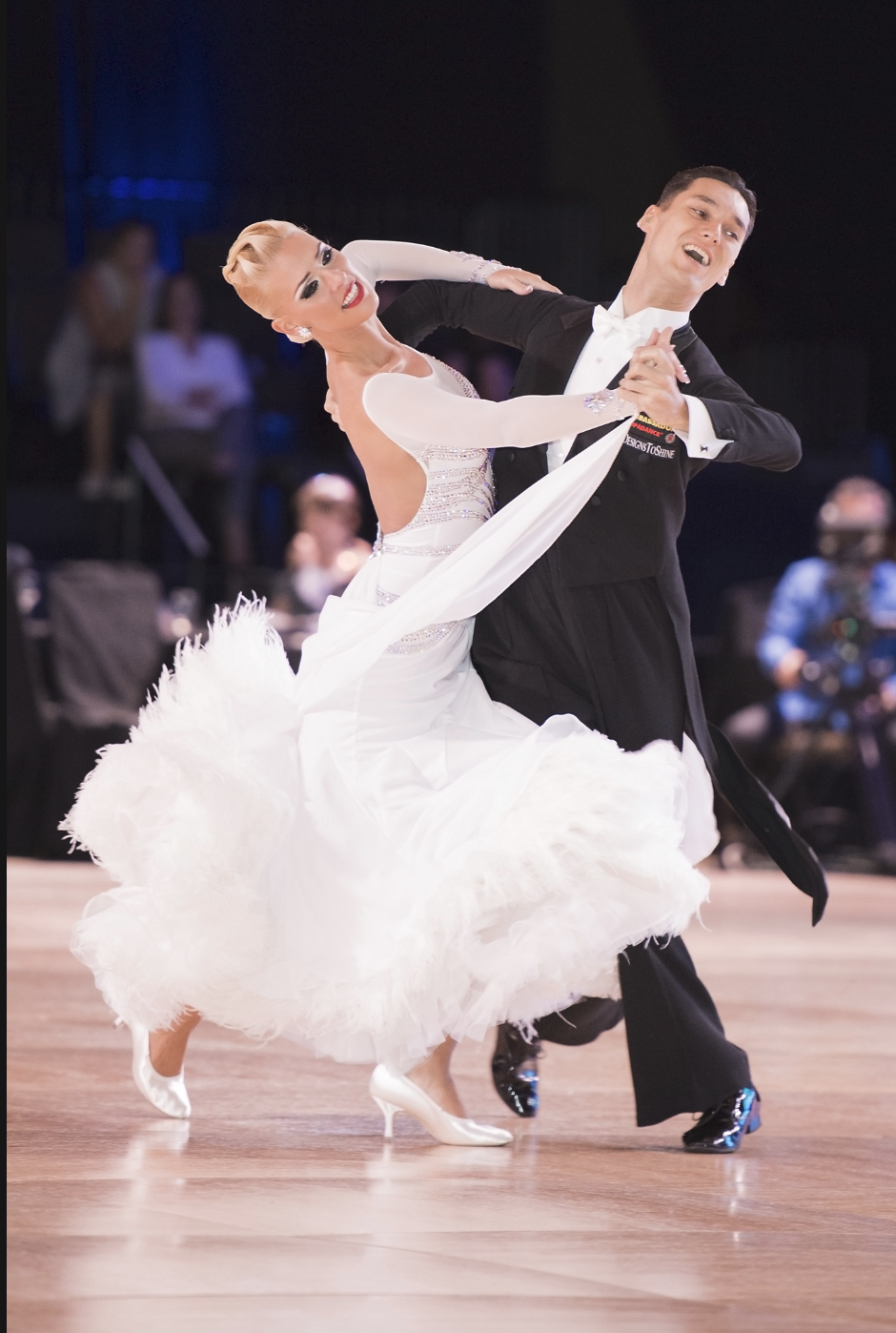 A man and woman dancing ballroom dance on a wooden floor, the woman in a flowing white gown and the man in a black tuxedo, with a blurred audience in the background.