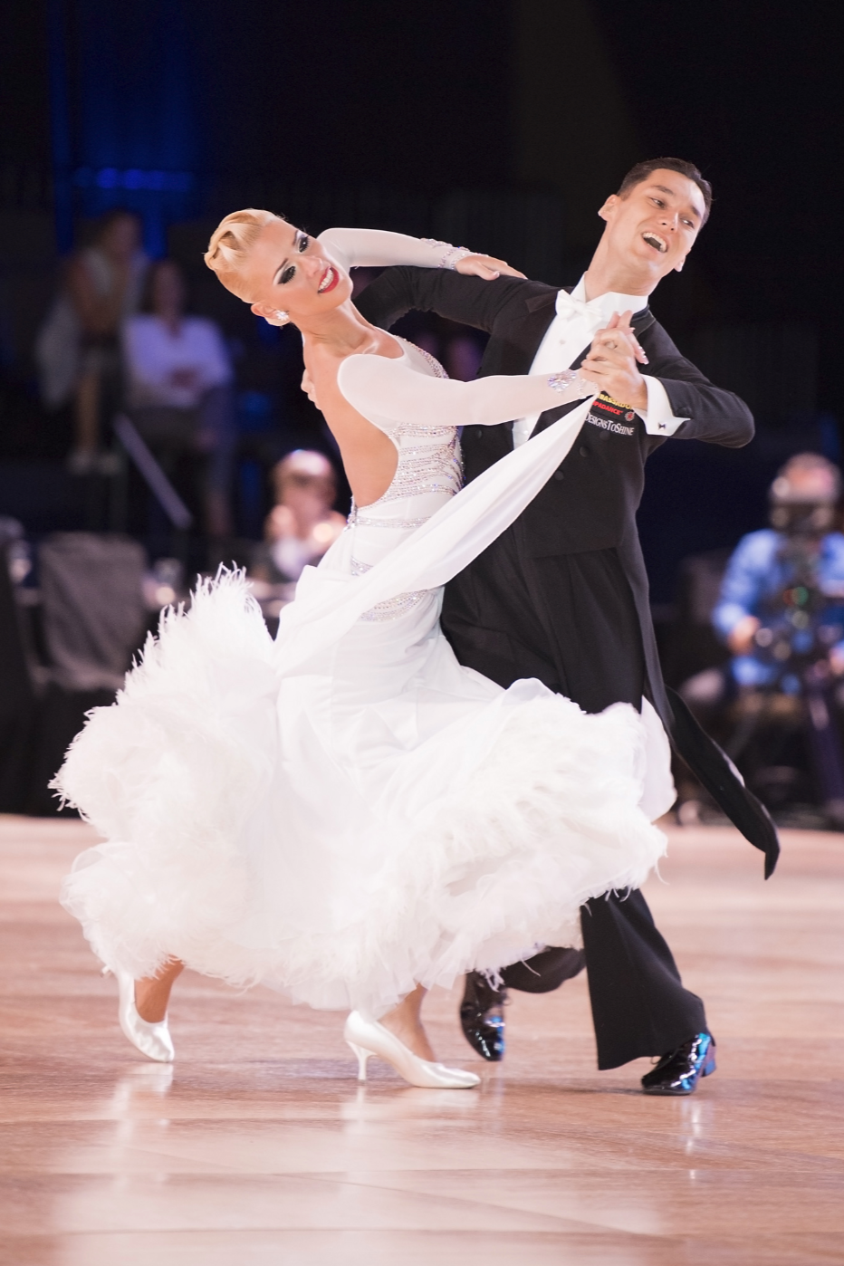 A couple performing a ballroom dance on a wooden floor, with the woman in a white gown and the man in a black tuxedo, surrounded by a blurred audience.