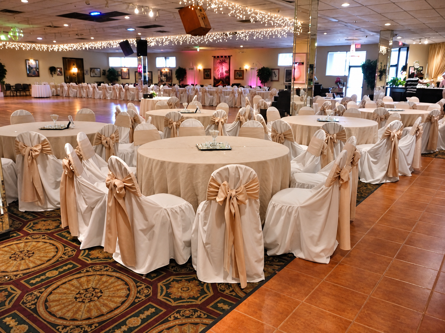 Event hall decorated with round tables covered in beige tablecloths. Chairs wrapped in white fabric with beige bow accents. String lights hanging from the ceiling, creating a festive atmosphere. An open space with a dance floor and a few framed pictures and plants along the walls.
